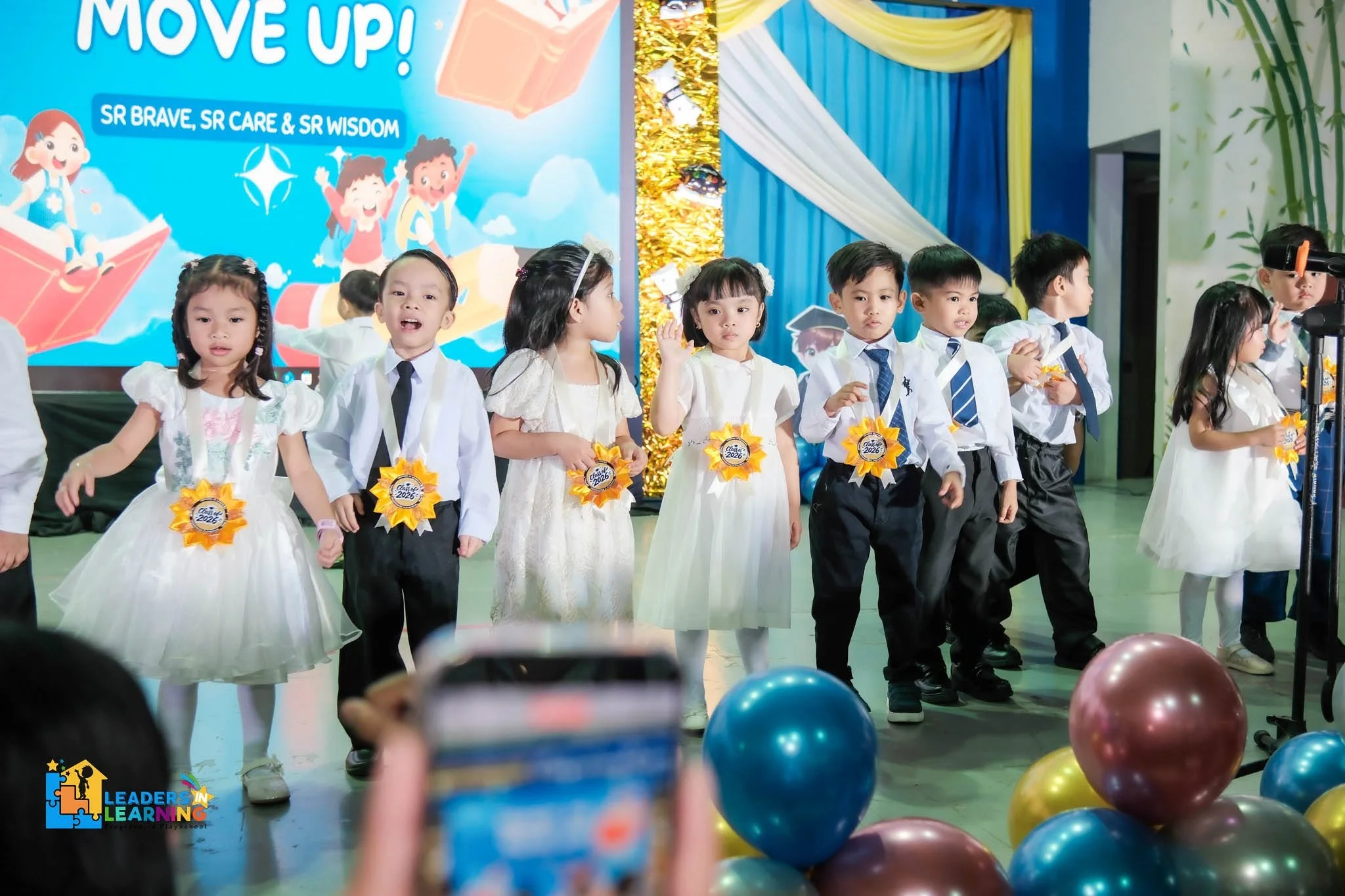 A group of young children dressed in formal clothes, some wearing ribbons that say 'Class of 2026,' are standing on a stage with colorful balloons in the foreground. The background features a large blue banner with cartoon characters and the words 'Move Up!' and 'SR Brave, SR Care & SR Wisdom,' indicating a graduation or moving-up ceremony for preschool or kindergarten students.
