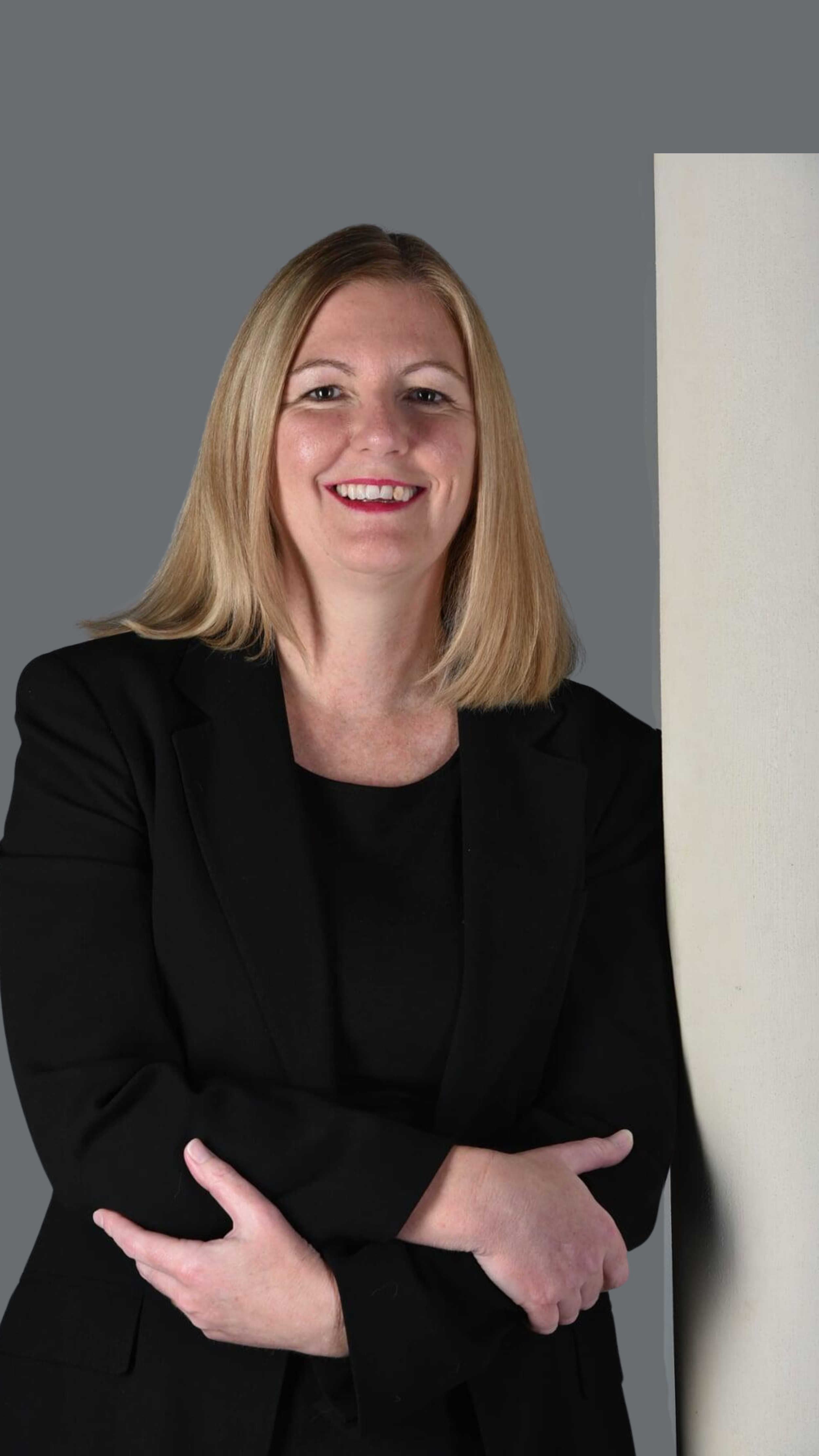 A woman with shoulder-length blonde hair, wearing a black blazer and top, smiling, standing against a plain grey background.