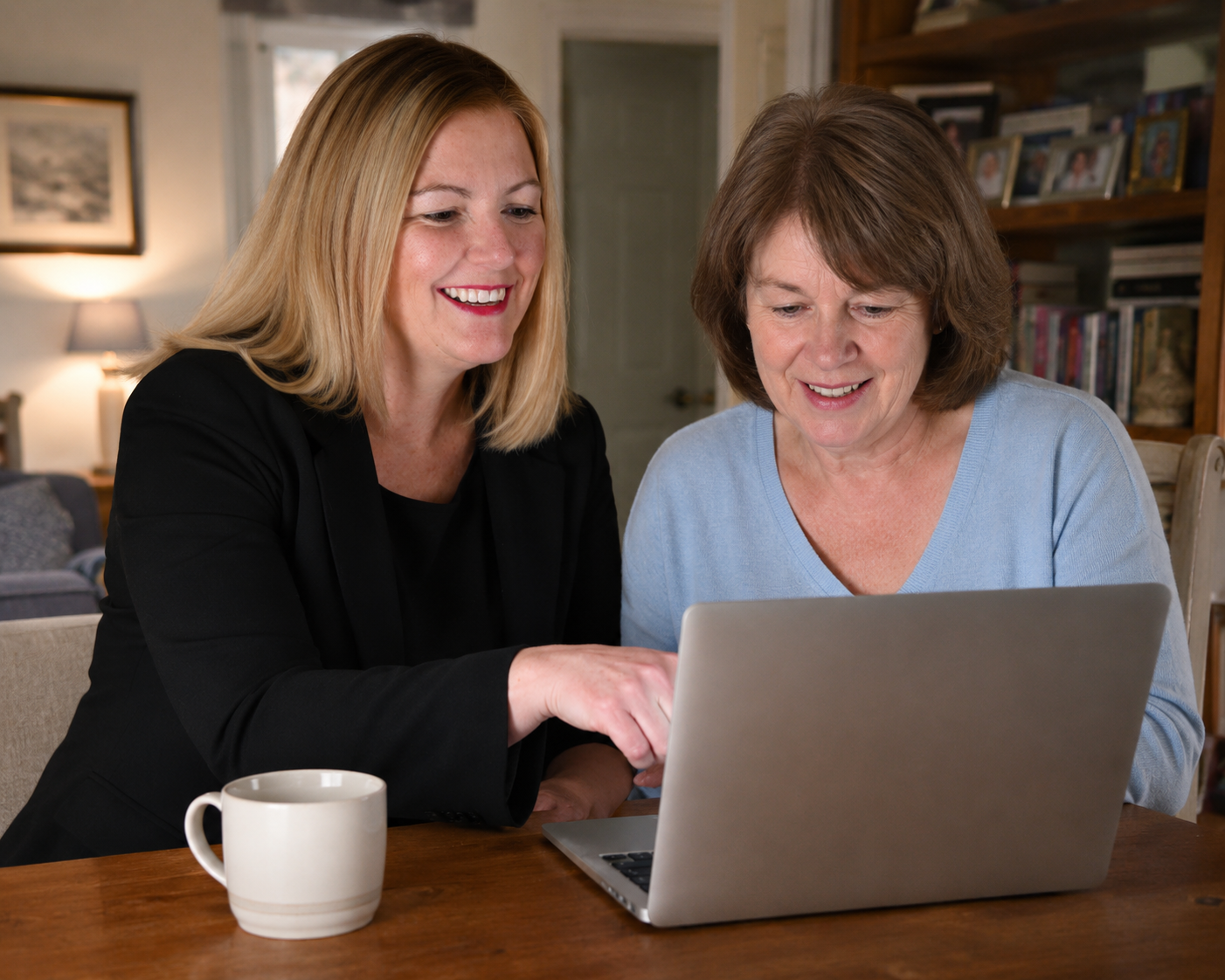 Two women sitting at a wooden table, looking at a laptop and smiling, with a white mug nearby.