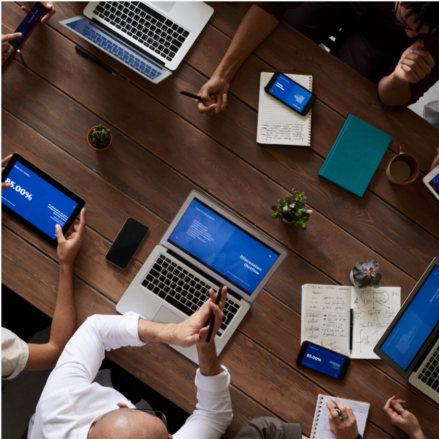 Overhead view of a wooden table with laptops, tablets, notebooks, a smartphone, a coffee mug, small potted plants, and people collaborating, some taking notes and others gesturing.