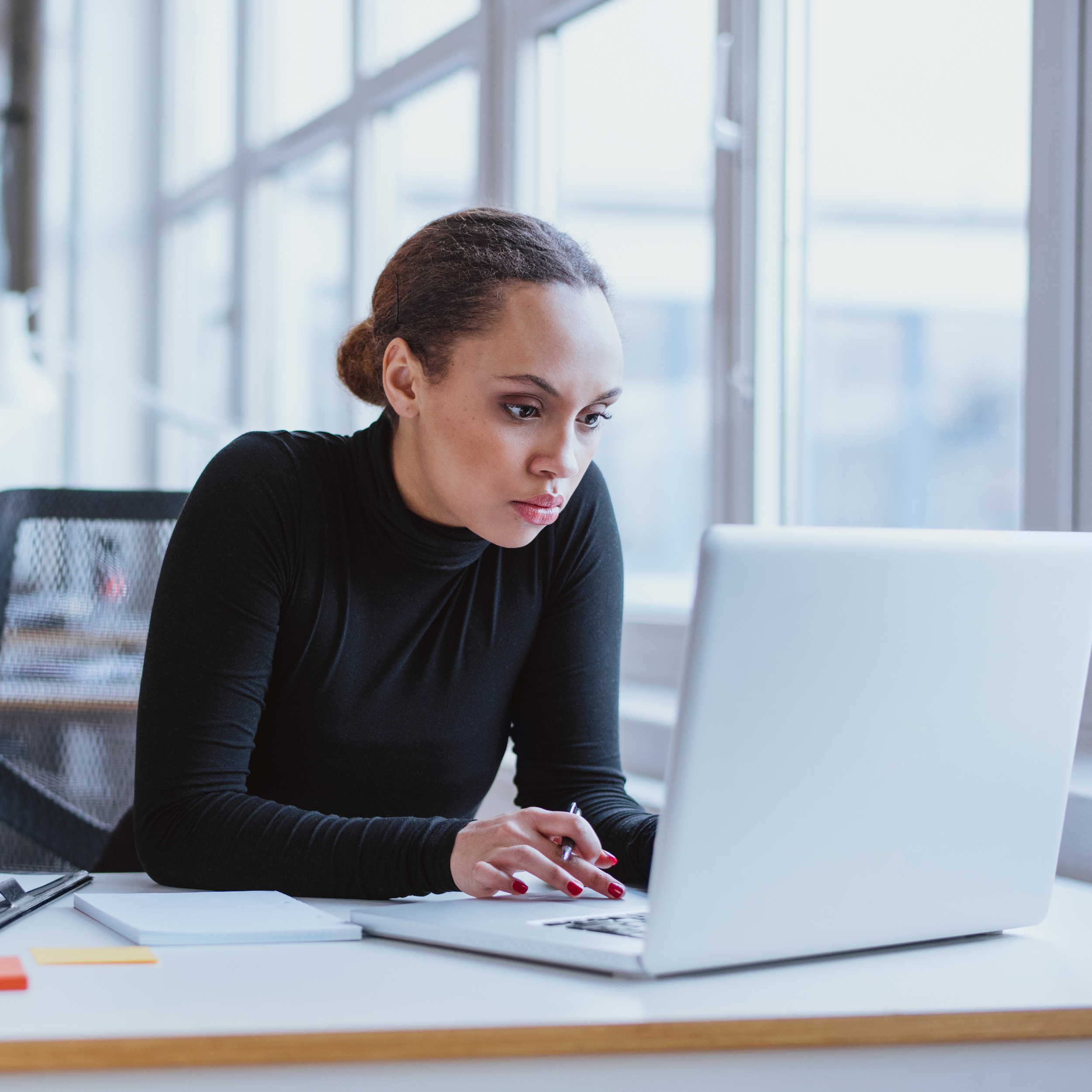A woman with dark hair tied back, wearing a black top, is sitting at a desk working on a laptop with a white surface. Behind her, there is a large window with a metal grid, allowing natural light to brighten the space.