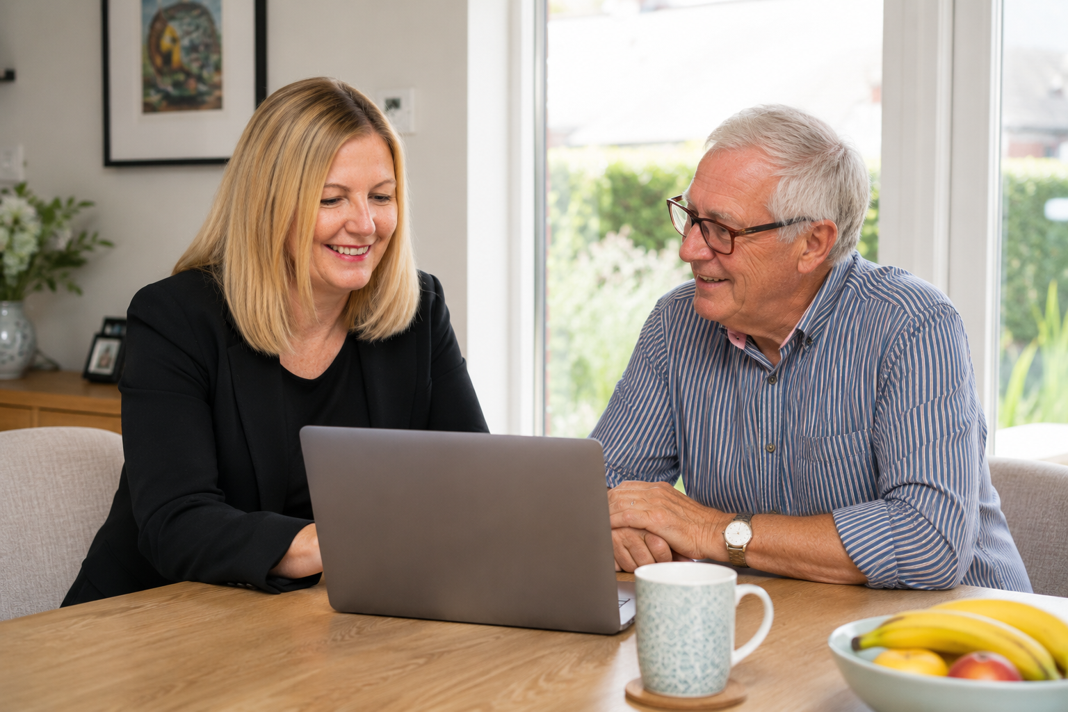 A woman and an older man sitting at a dining table, looking at a laptop and smiling, with a bowl of bananas and apples, a coffee mug, and a window with outdoor greenery in the background.