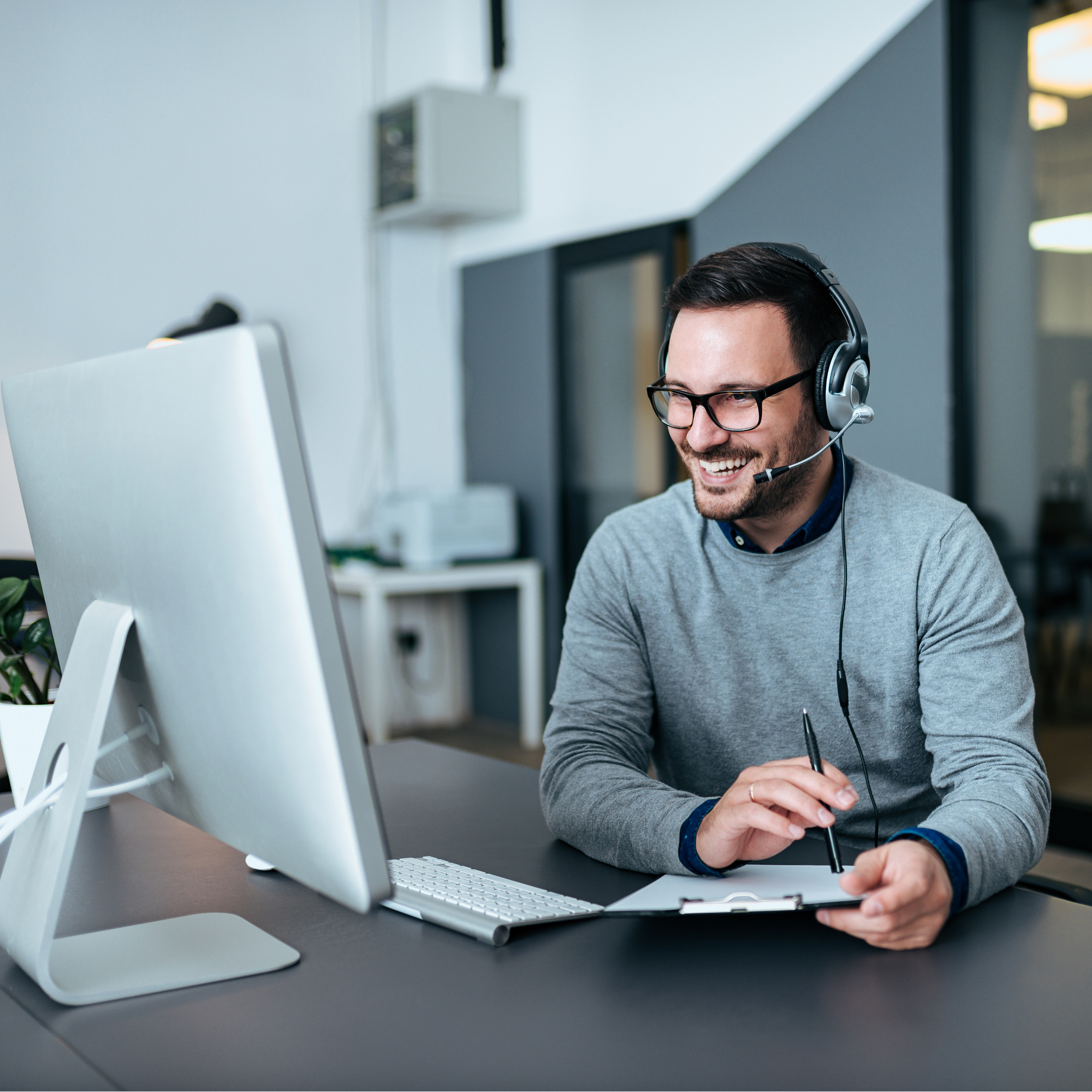 A man wearing glasses and a headset smiling while working at a computer in a modern office.