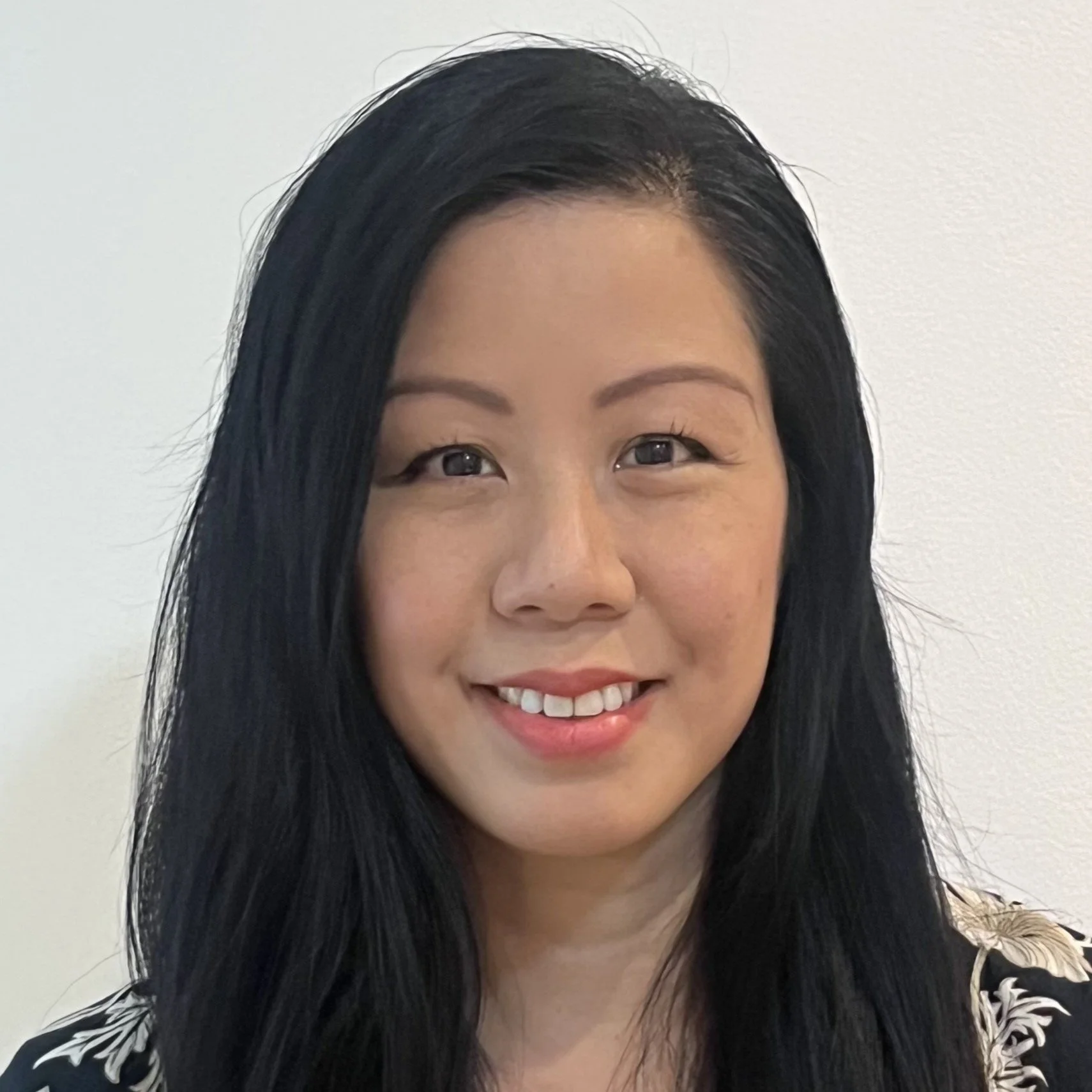 A woman with long black hair smiling in front of a plain white wall.