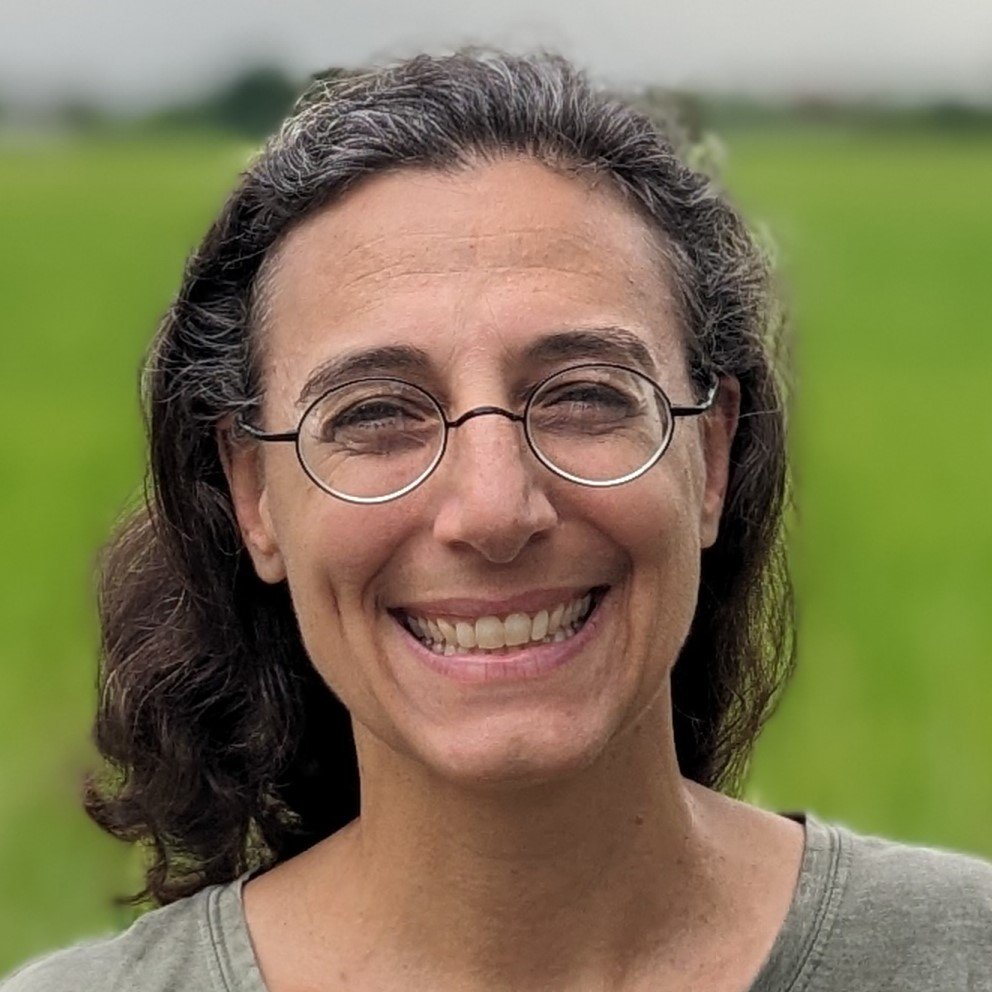 A smiling woman with curly brown hair and round glasses standing outdoors with a green field in the background.