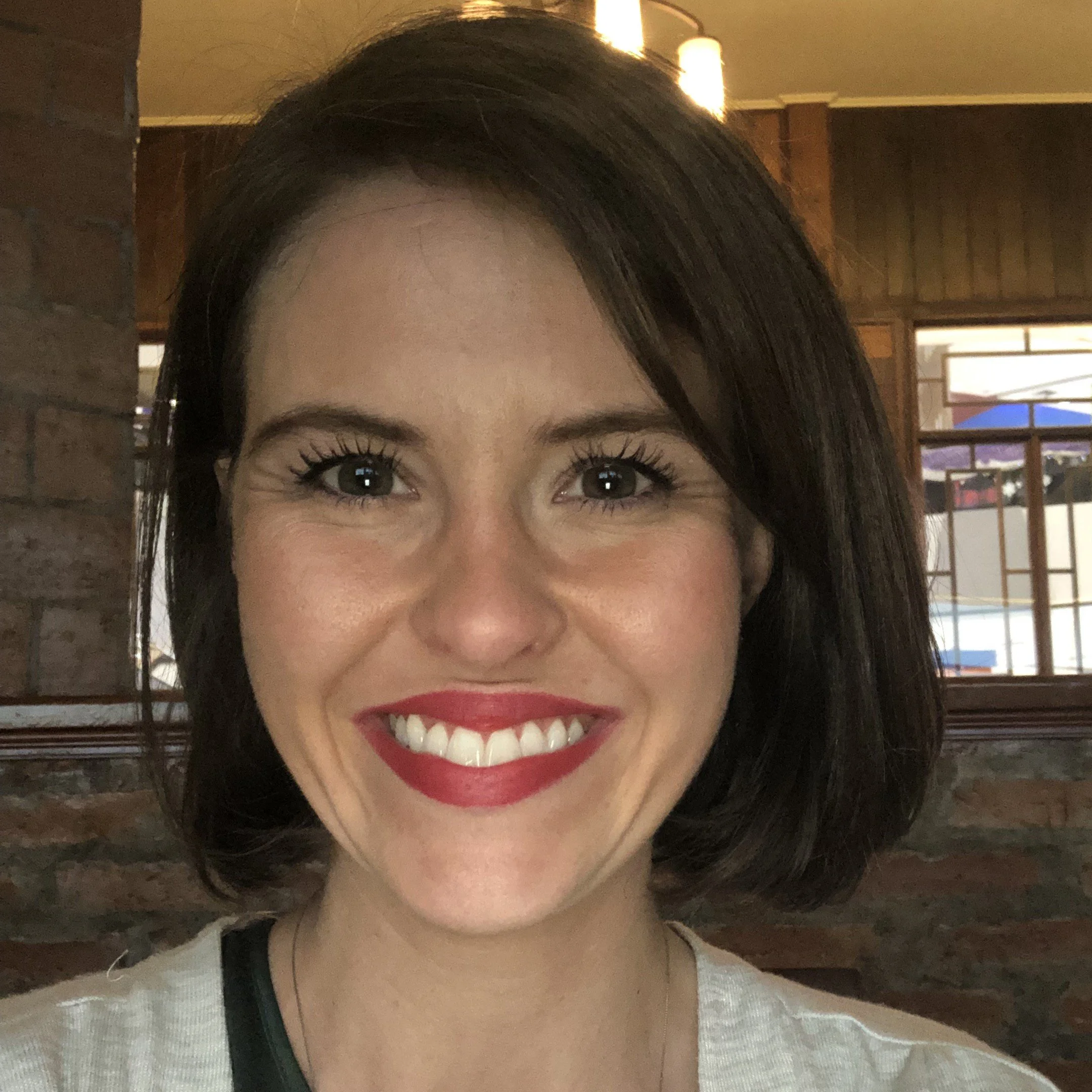 Close-up of a smiling woman with dark hair, wearing red lipstick and a white top, indoors with wooden walls and windows in the background.