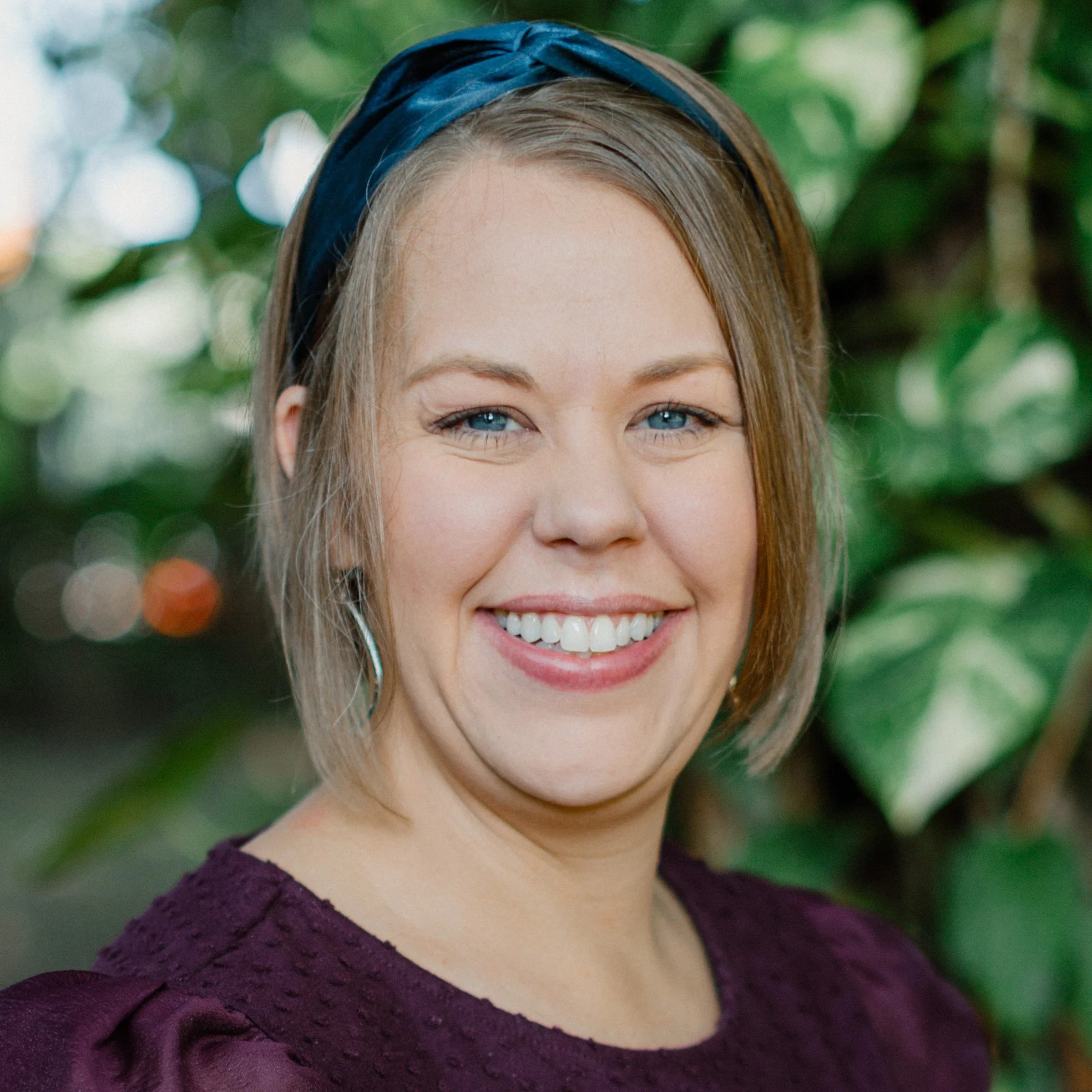 A woman with blue eyes, blonde hair, and a wide smile, wearing a purple dress and a blue headband, outdoors with green foliage in the background.
