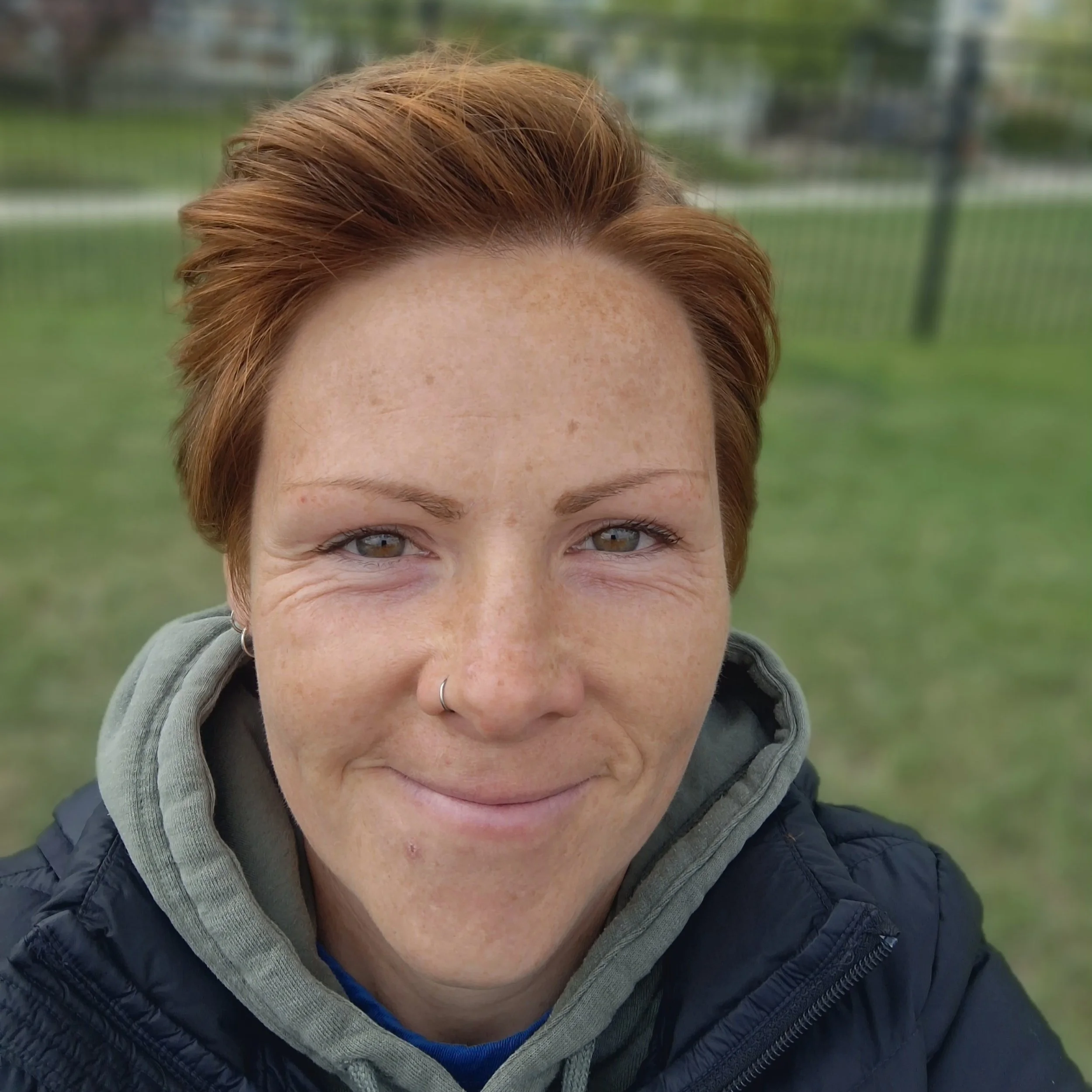 Close-up of a person's face with short, reddish hair, freckles, and blue eyes, wearing a gray hoodie and a black puffer jacket, outdoors with green grass and trees in the background.