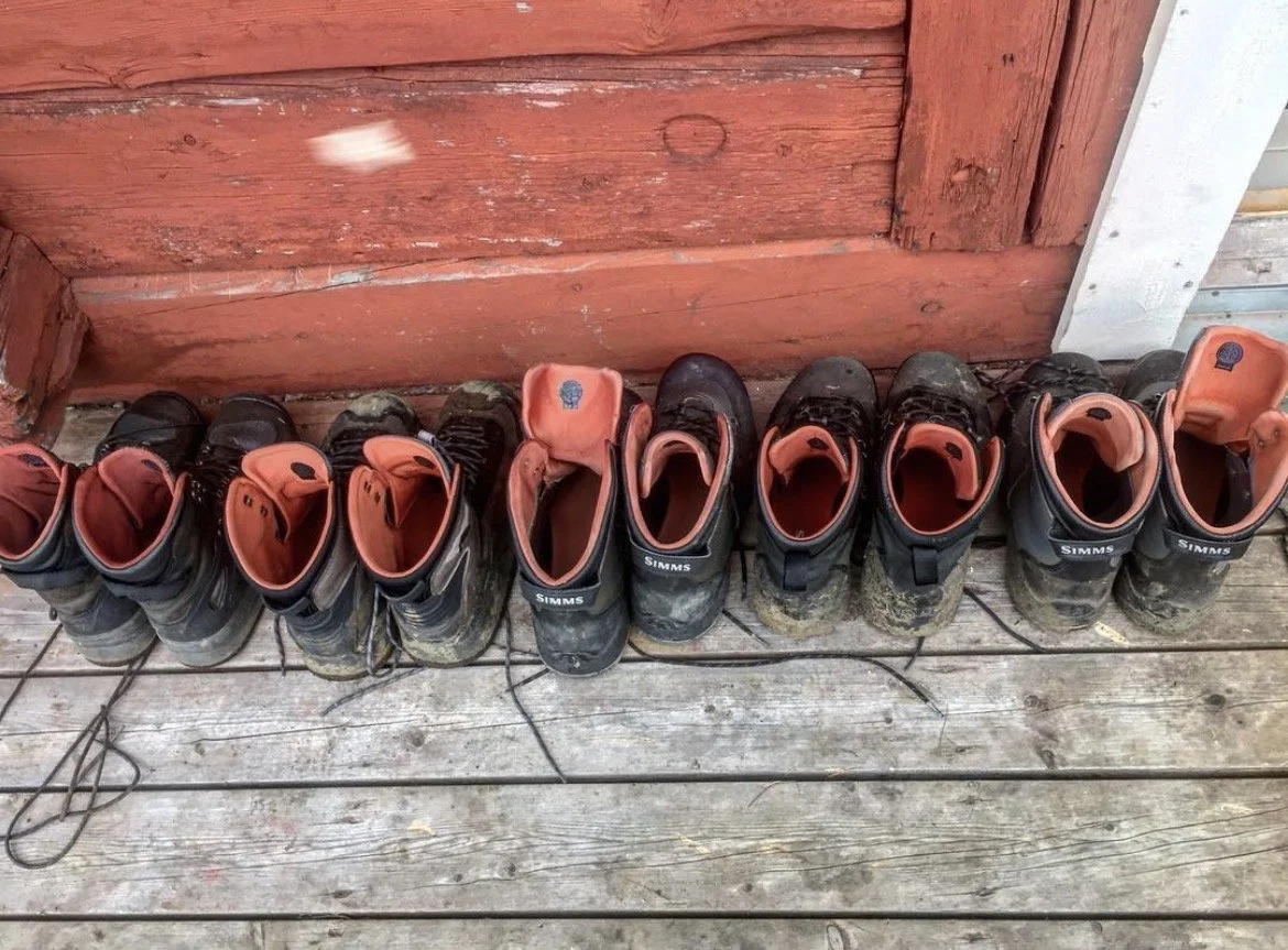 A row of muddy work boots lined up on a wooden porch in front of a red wooden wall.