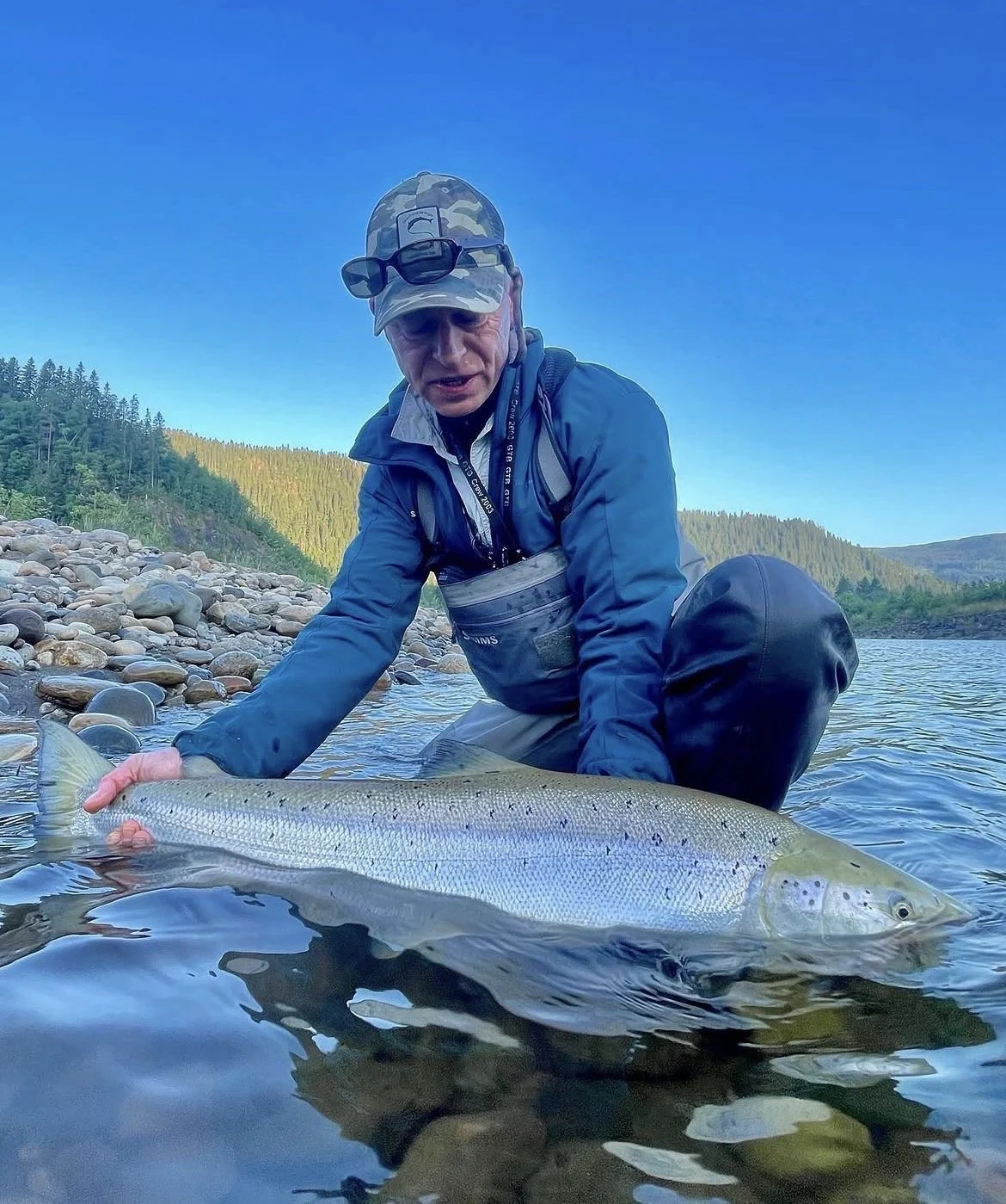 A man wearing outdoor gear and a camouflage cap kneels in a river, holding a large fish he caught during a fishing trip with hillside and forest in the background.
