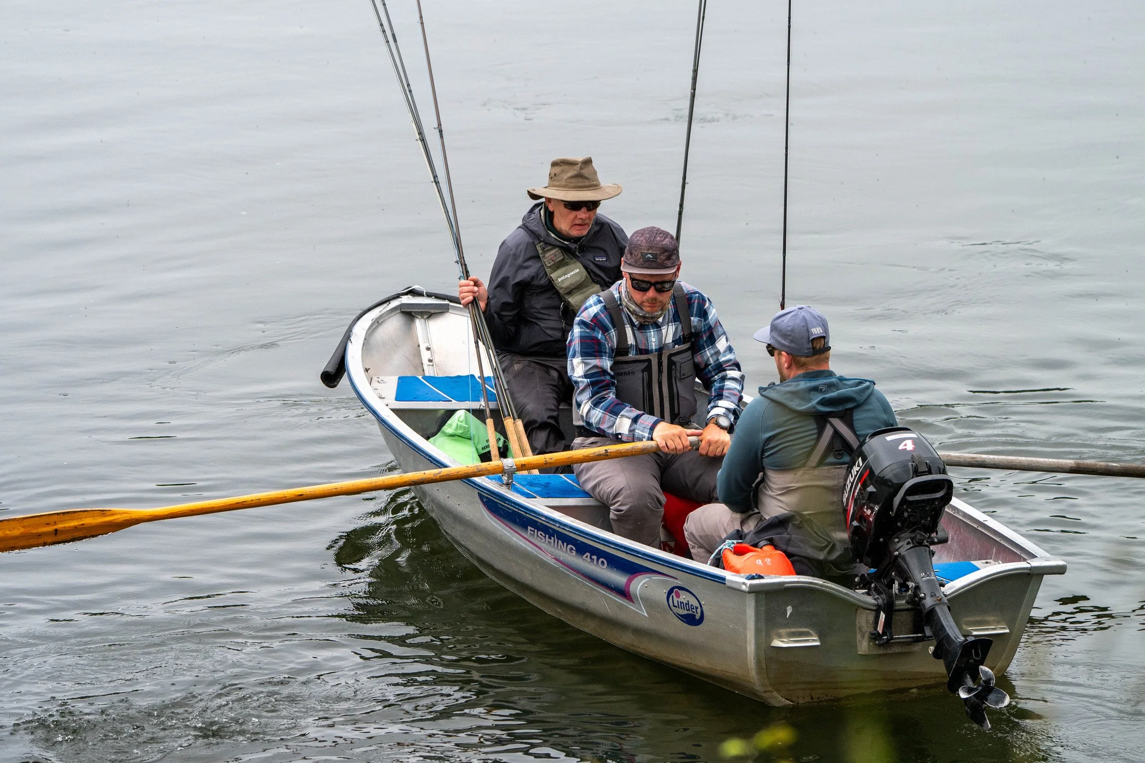 Three men in a small boat on a body of water, preparing to fish, with fishing rods and an outboard motor.