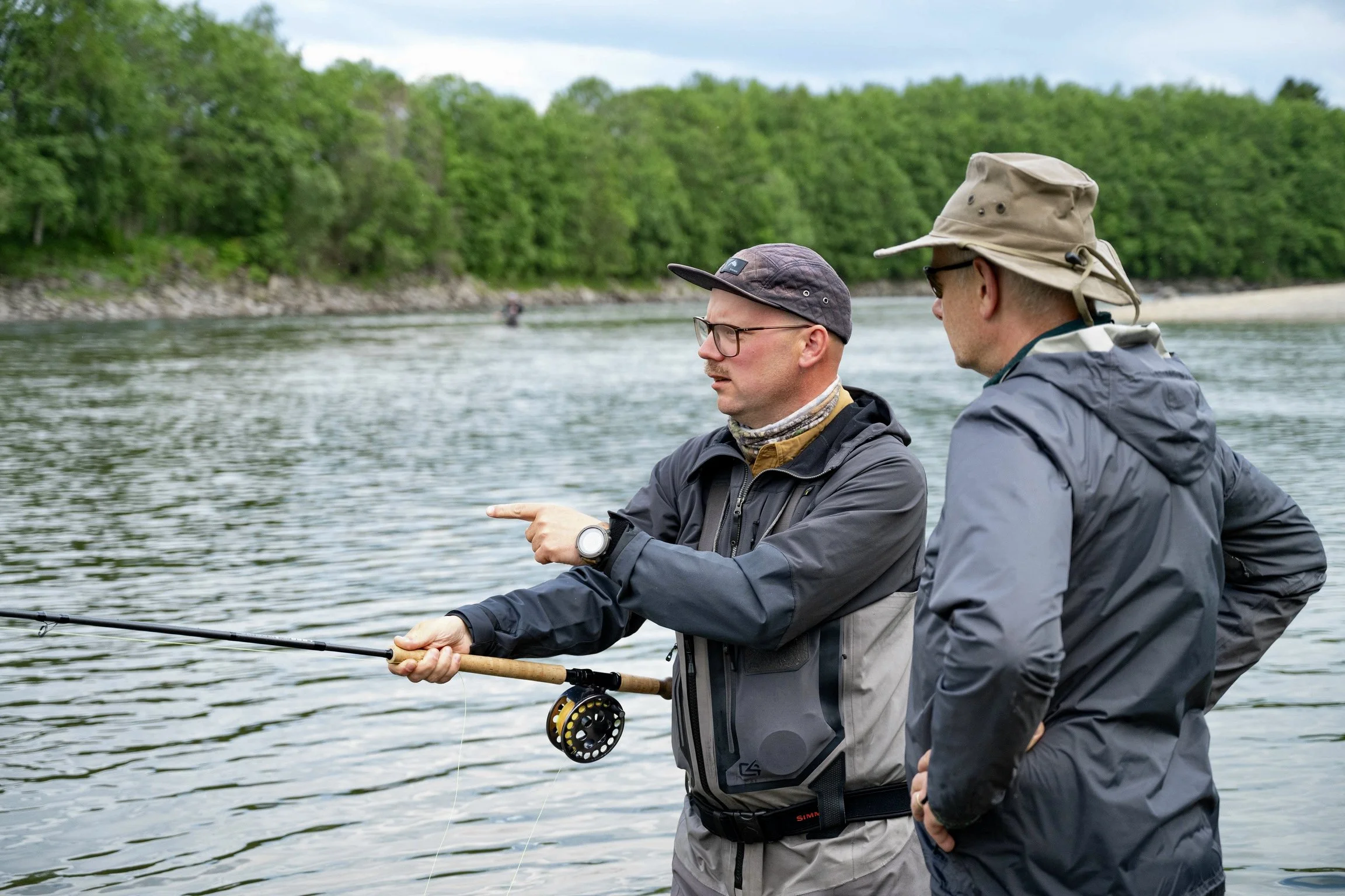 Two men fishing in a river, surrounded by lush green trees, with one man pointing at his fishing rod.