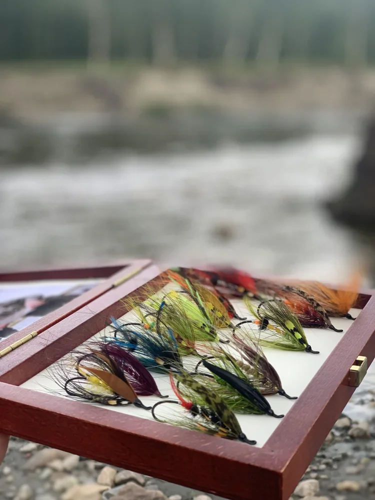 A wooden box with colorful fishing flies placed inside, set outdoors near a river with rocks and trees in the background.
