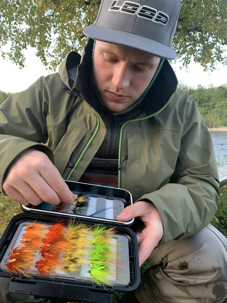 A man outdoors near water, wearing a gray cap with 'Leep' logo, olive jacket, and fishing gear, sorting a collection of colorful fishing flies in a box.