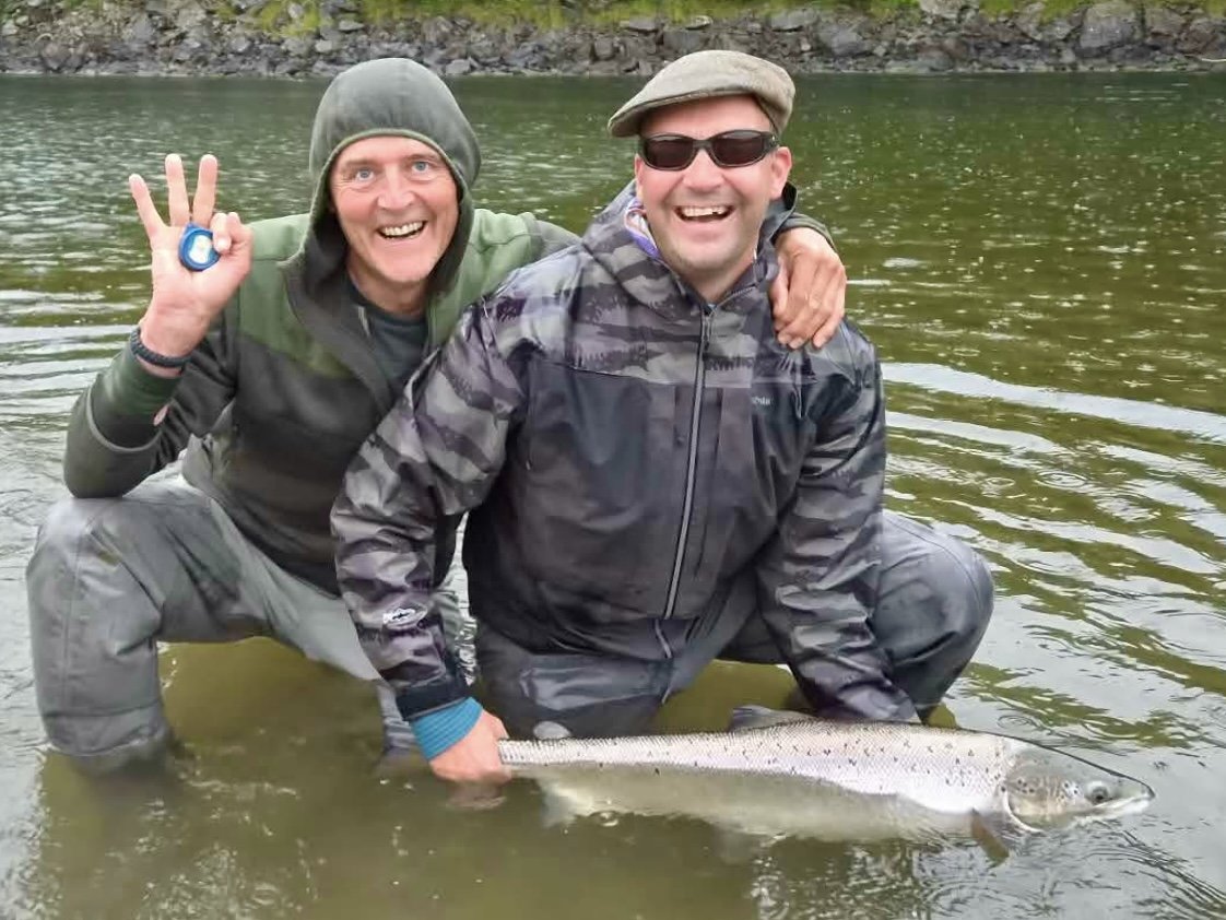 Two men fishing in a river, holding a large fish, smiling, with one giving a victory sign, dressed in outdoor gear and hats.