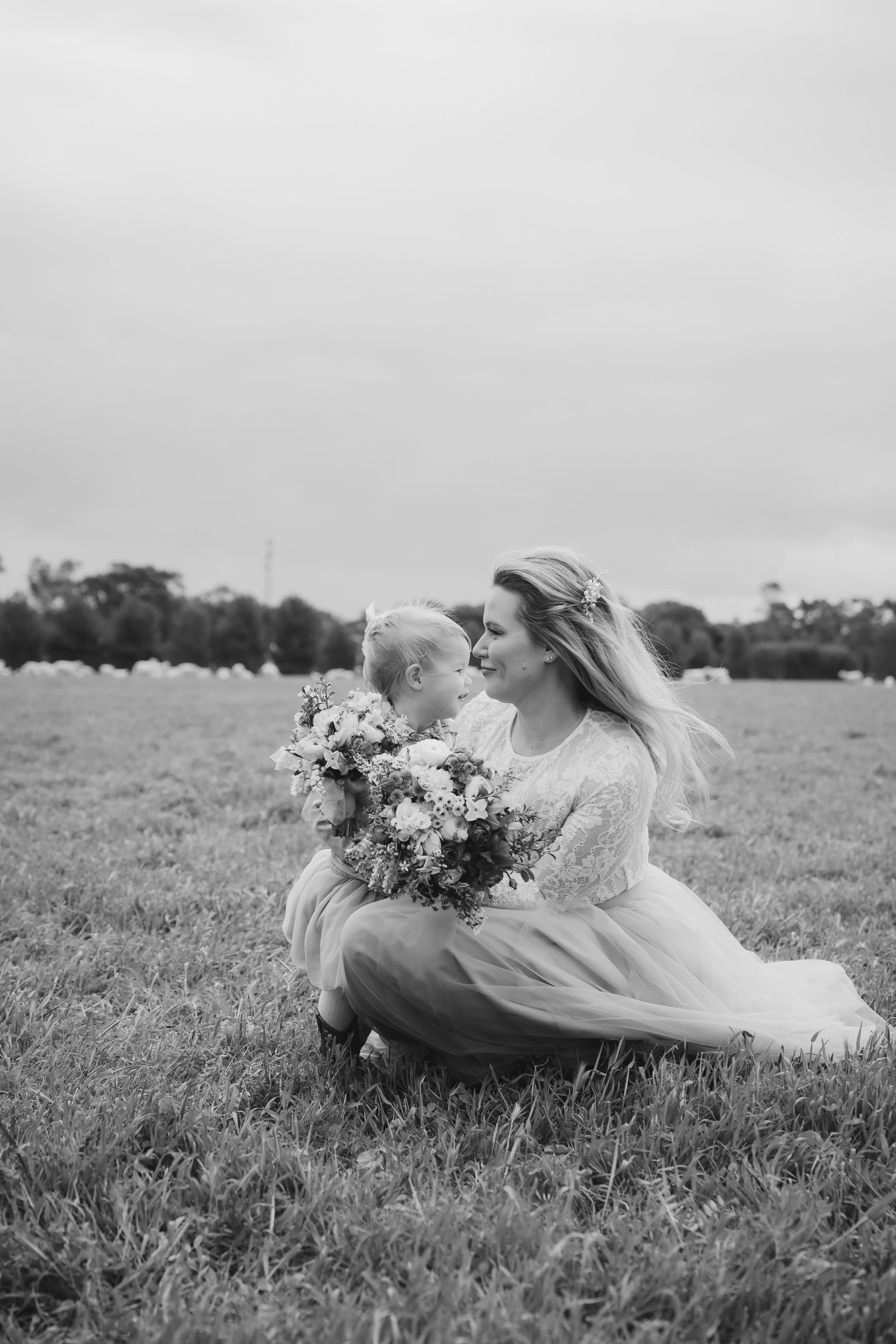 A woman in a wedding dress kneeling on grass holds a young girl in a dress and glasses. They are face to face, smiling and holding a bouquet of flowers, with trees and a cloudy sky in the background.