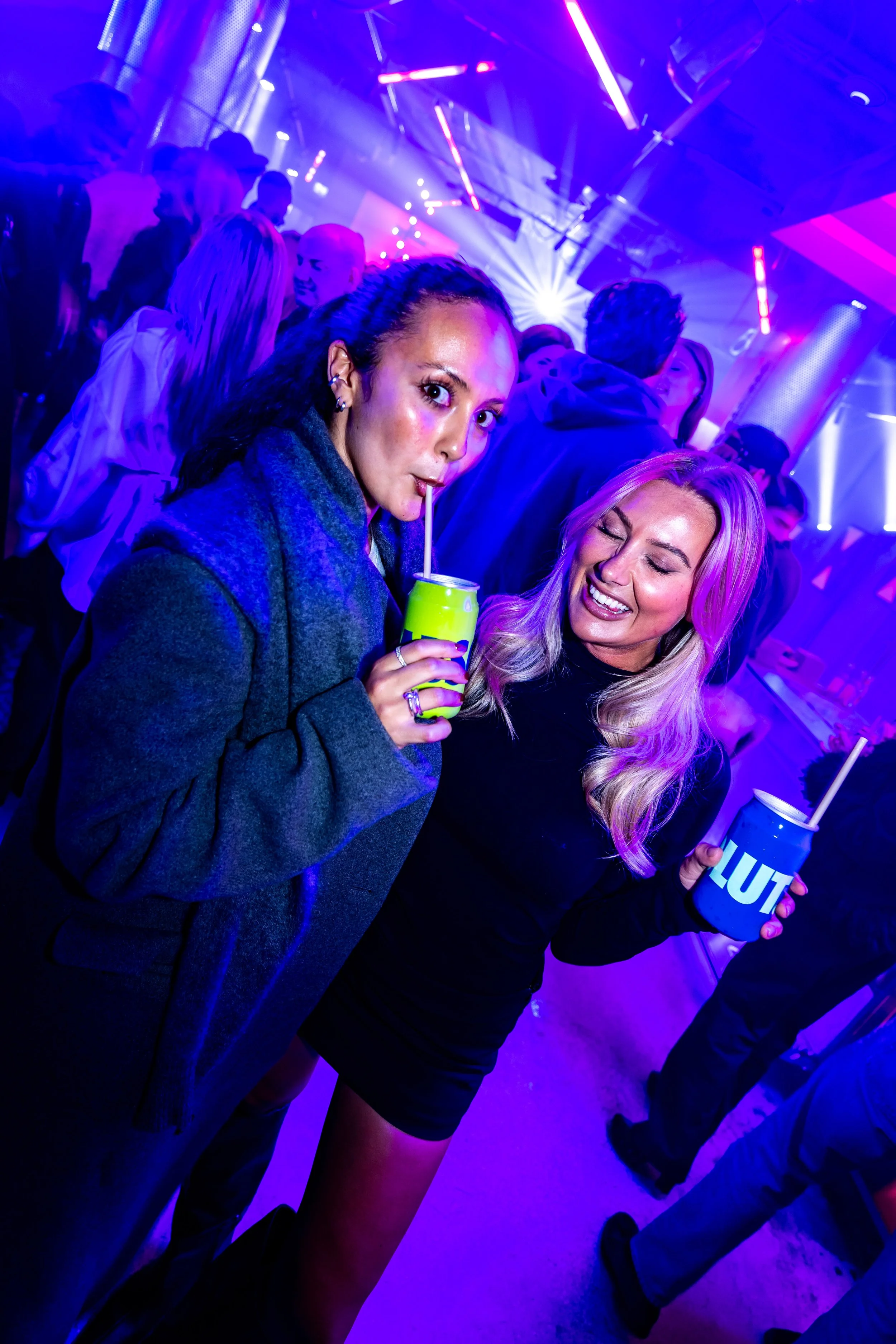 Två kvinnor som tycker om att dricka drycker på ett nattklubb med färgstarka neonljus i bakgrunden, omgivna av andra människor.