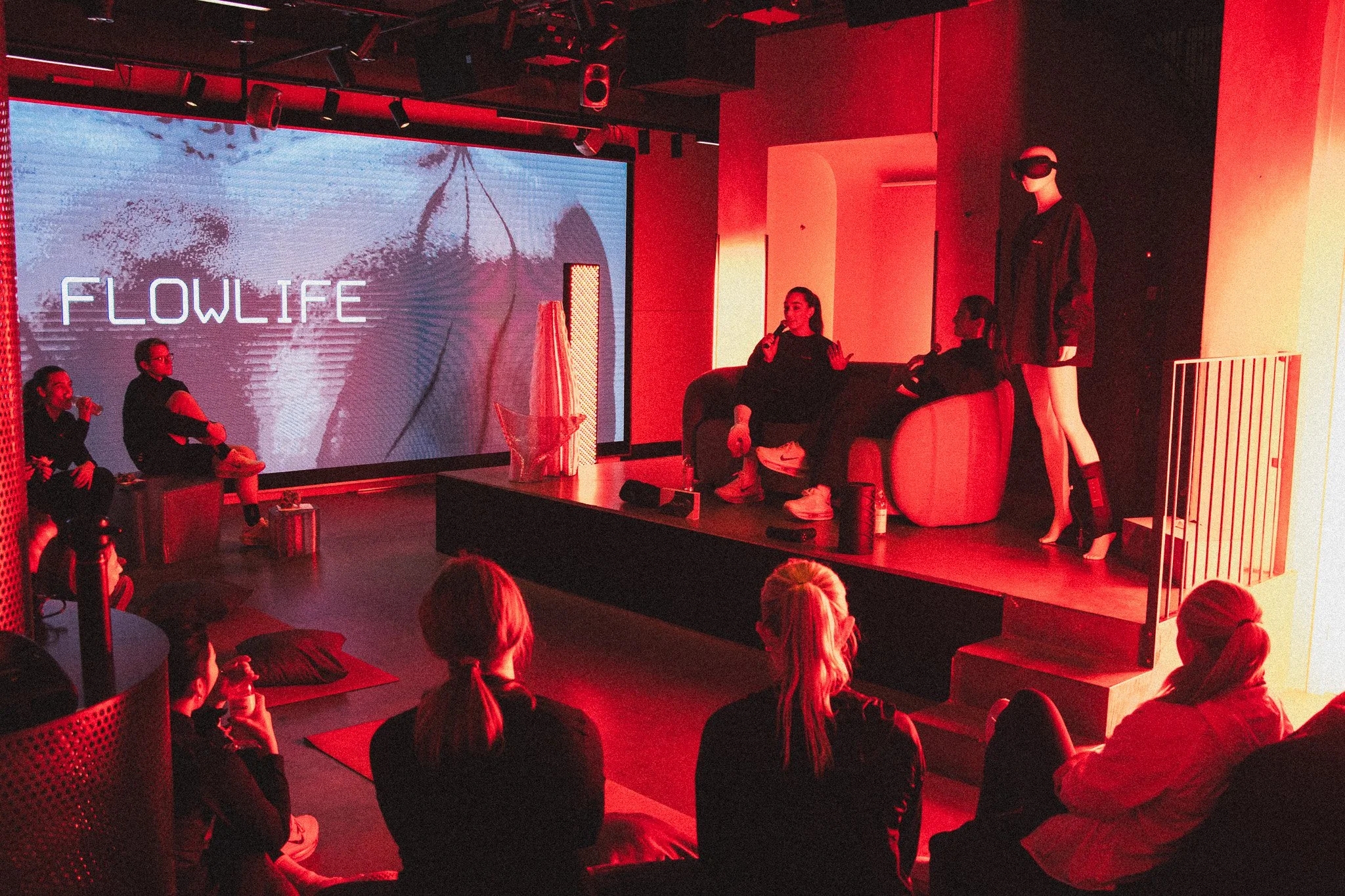 En grupp människor som sitter och lyssnar på en presentation i ett mörkt rum med röda belysningar, en stor skärm med texten "FLOWLIFE" visas på bakgrunden.