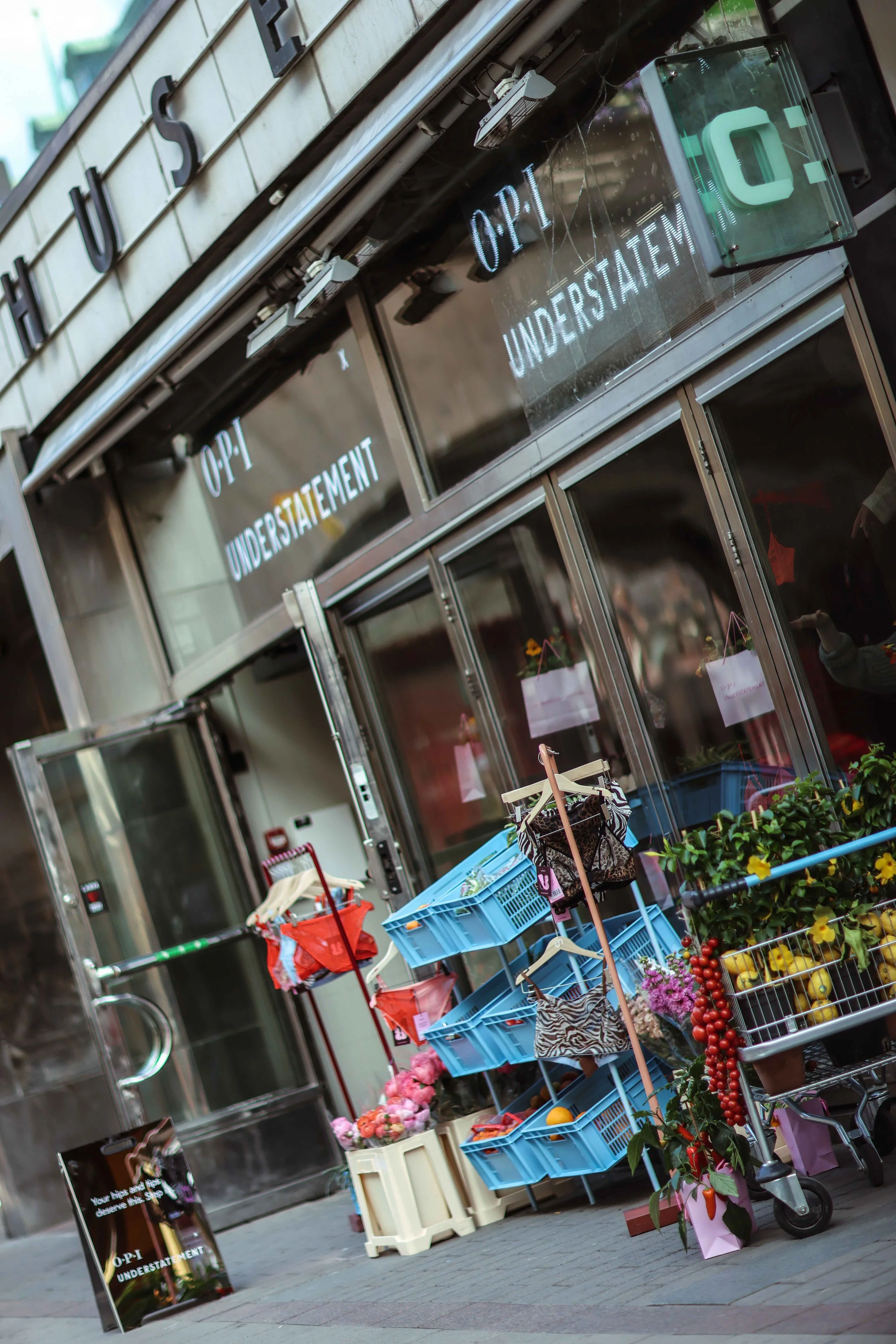 Utanför en butik finns en small karusell med blomsterarrangemang, växter och frukt för försäljning. Det finns också en skylt med texten 'OPI UNDERSTÄTEMENT'.