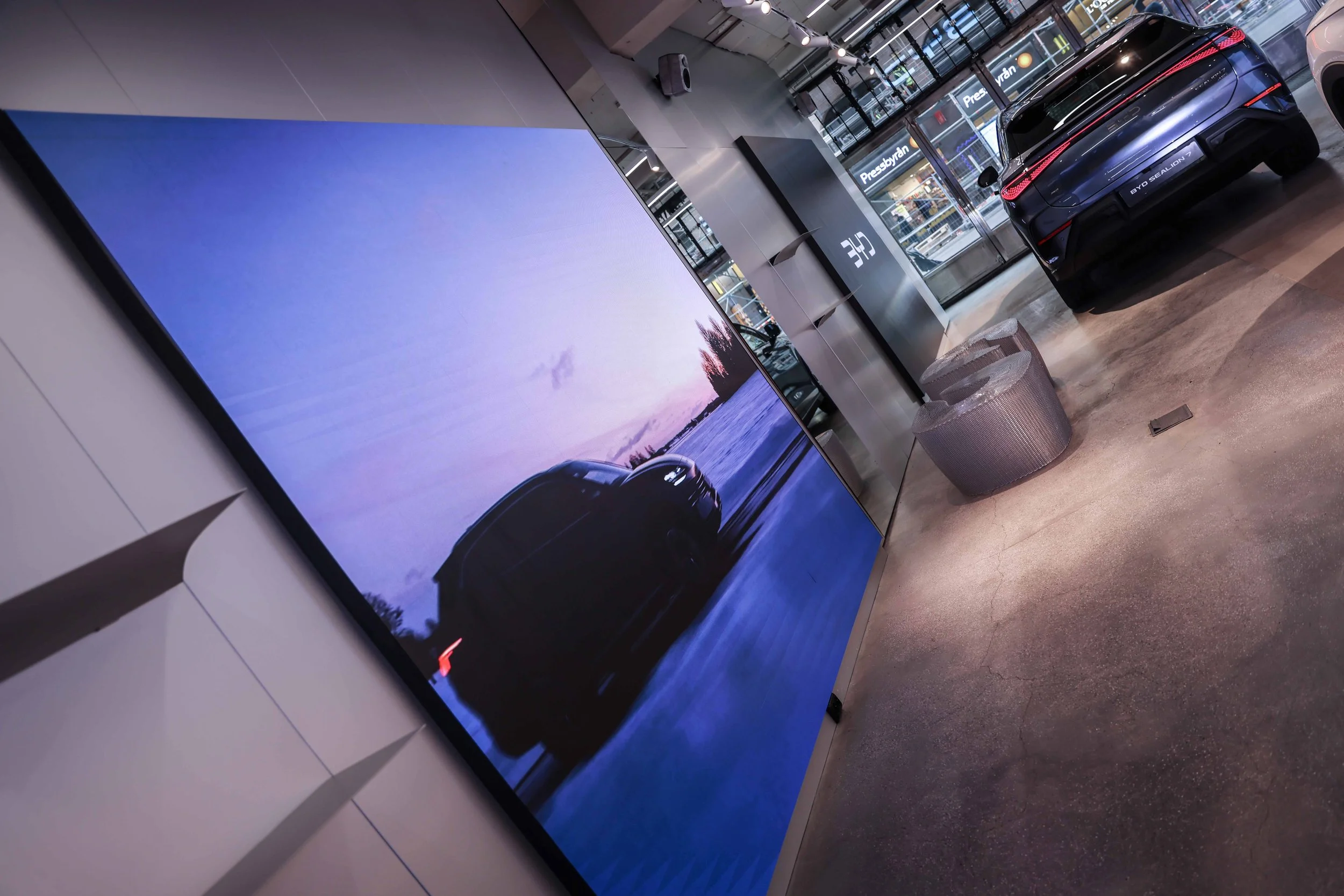 En stor digital skärm i en modern utställningshall visar en bil vid en vacker solnedgång vid vattnet. Bilen är en svart BYD Sealion 7.