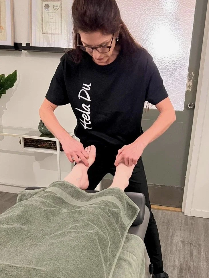 En kvinna ger en fotmassage eller behandling till en person som ligger på en massagebänk, i ett rum med medicinska eller terapeutiska certifikat på väggarna.