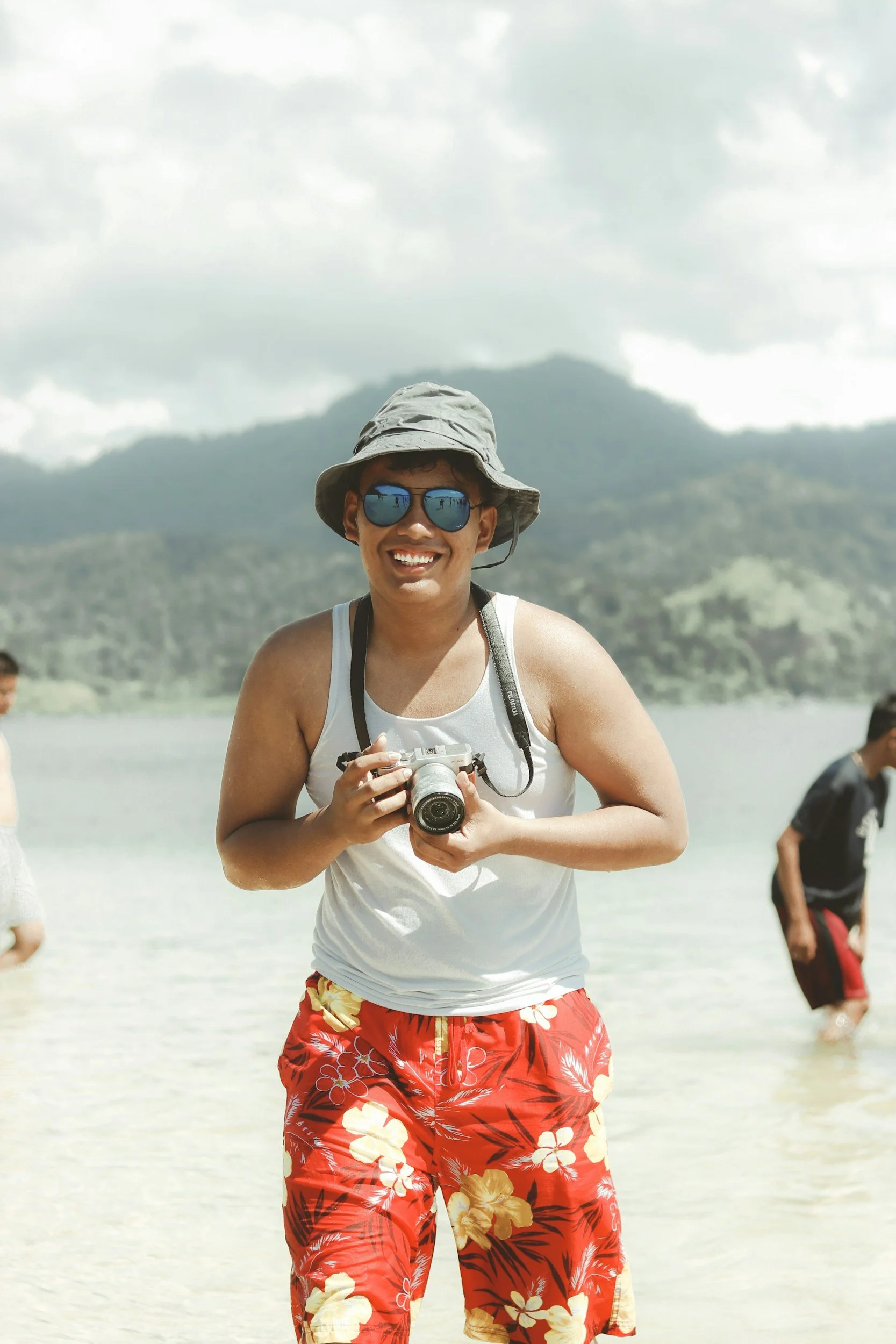 Young man in sunglasses, bucket hat, white tank top, and red floral shorts holding a camera, standing on the beach with cloudy sky and mountains in the background.
