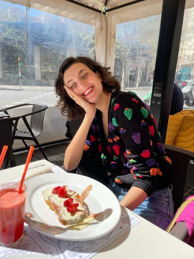 Una joven sonriendo con los ojos cerrados, posando en una cafetería, con un pastel de frutas y una bebida en la mesa.