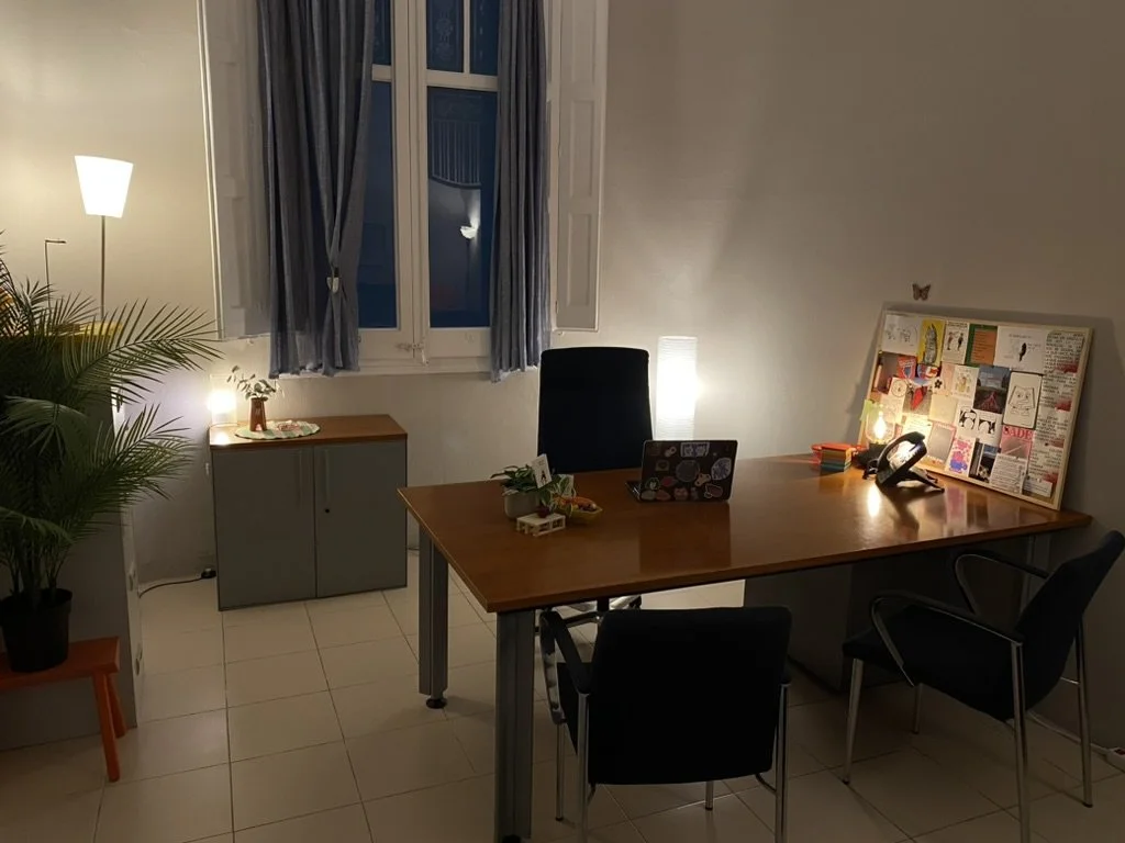 Despacho con escritorio de madera, silla de oficina, computadora portátil decorada con pegatinas, lámpara de pie, ventanas con cortinas azules y plantas de interior