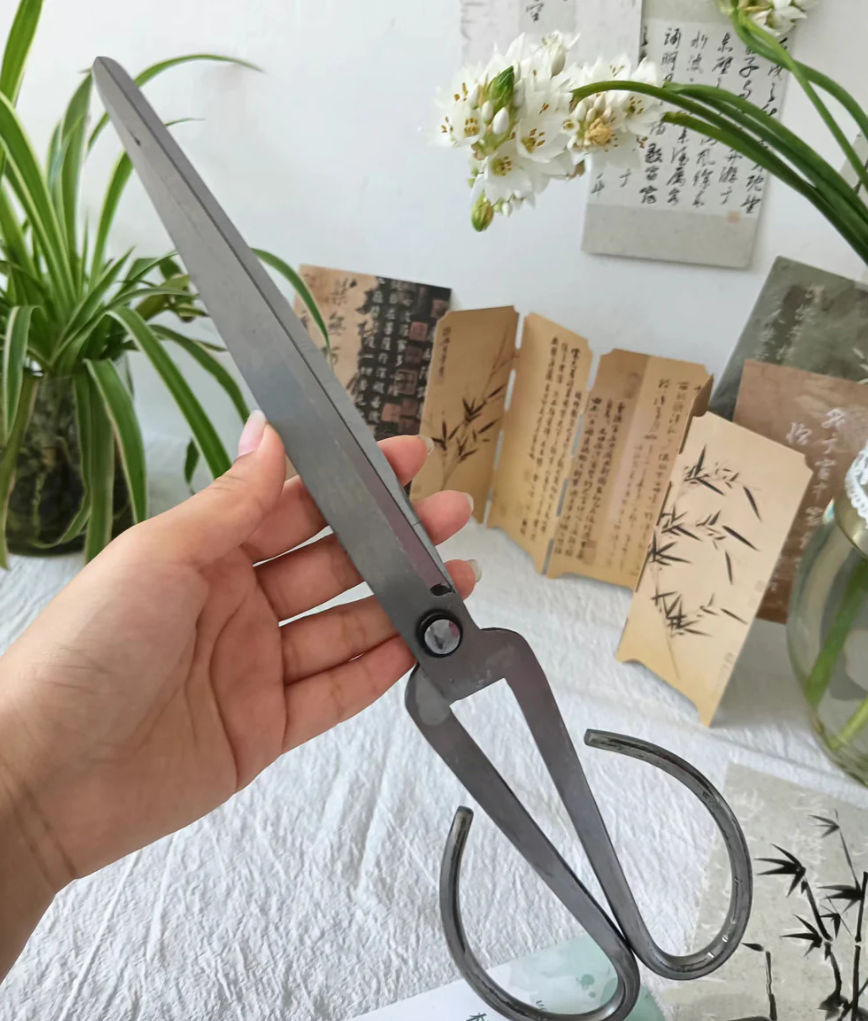 Person holding a pair of large metal tongs or scissors near a table with botanical and Asian-style paper decorations, with a plant and flowers in the background.