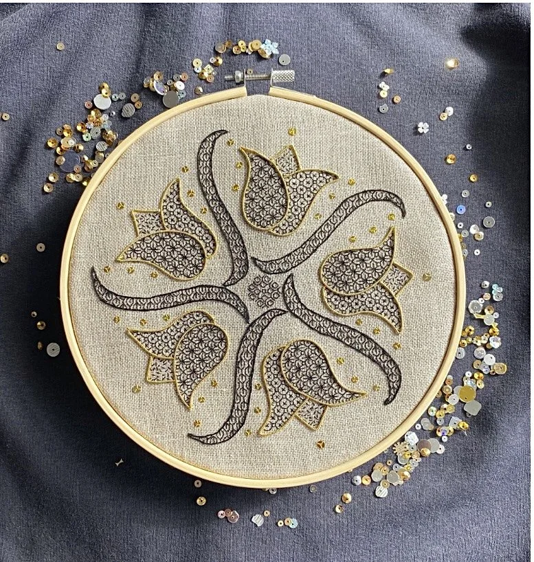Embroidery hoop with intricate black and gold floral design on beige fabric, surrounded by scattered sequins on dark fabric background.