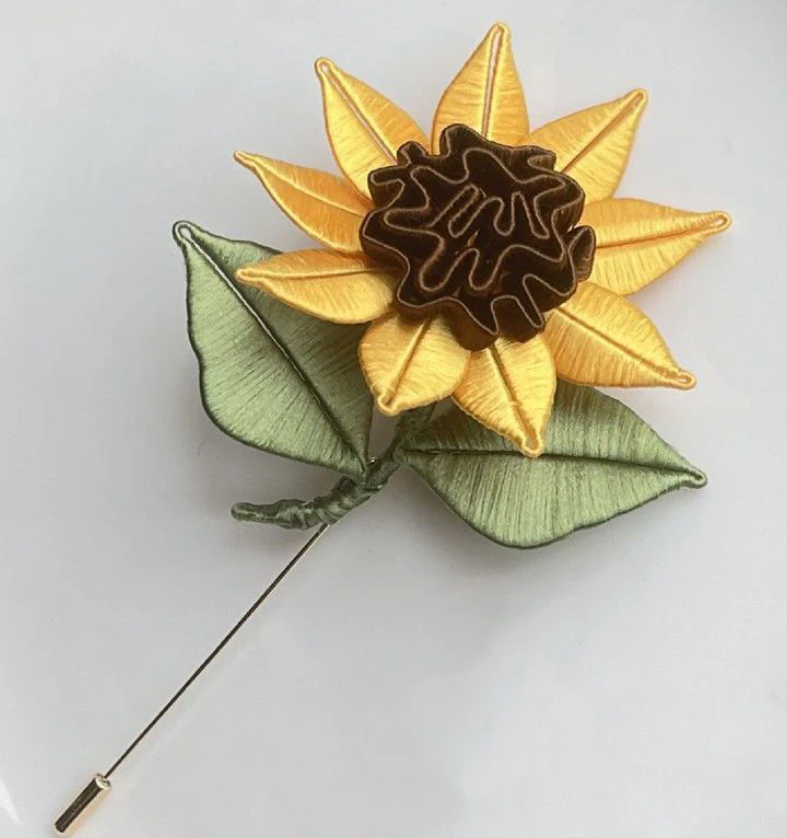A decorative brooch shaped like a sunflower with yellow petals and green leaves, featuring a dark center pressed against a plain white background.