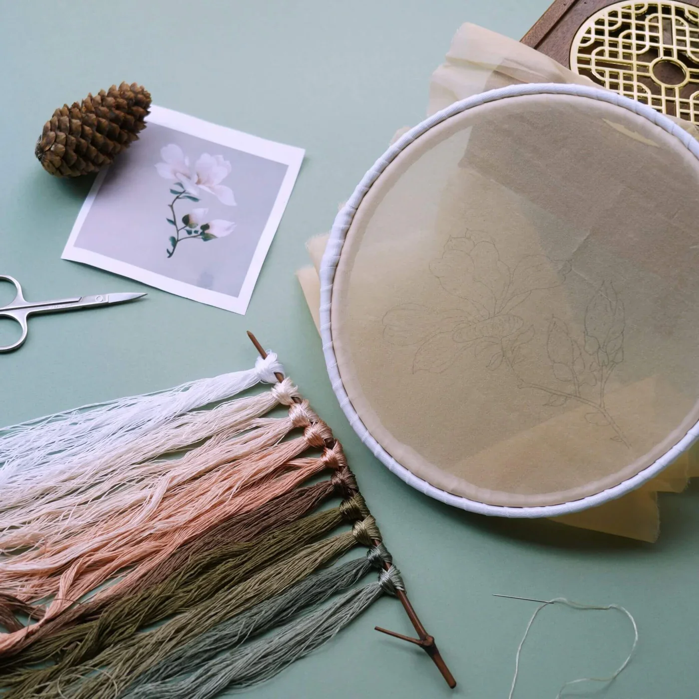 Embroidery hoop with fabric, sketch of floral design, color sample tassels, pinecone, scissors, thread, and a printed photo of a flower on a light green surface.