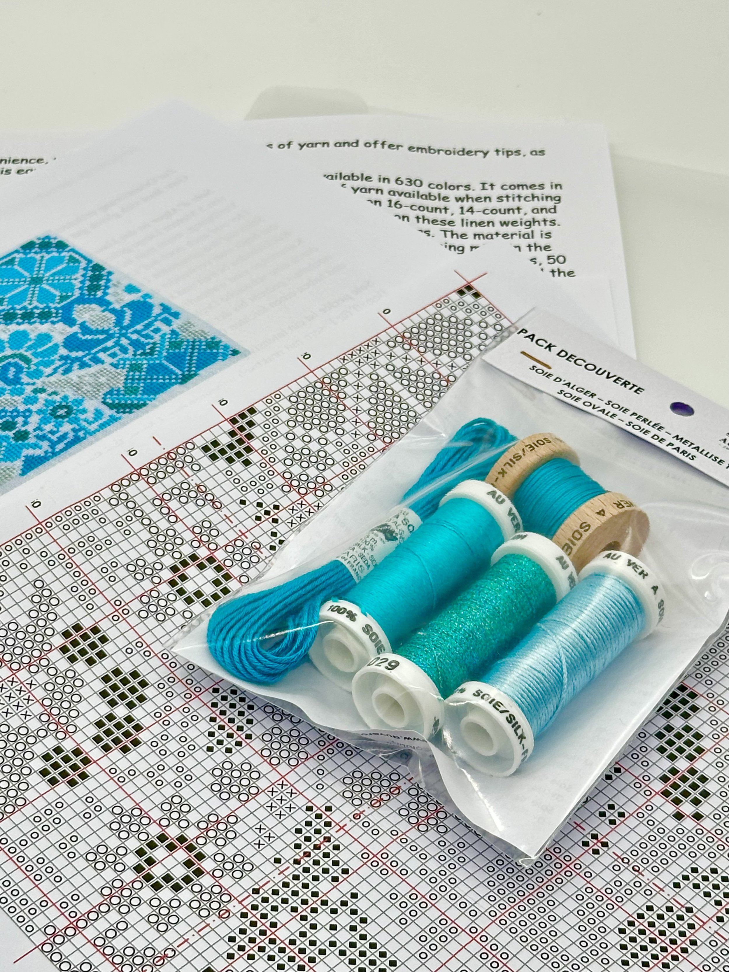 A collection of blue embroidery floss spools on clear plastic packaging, resting on a cross-stitch pattern leaflet with blue and black thread samples and design charts.