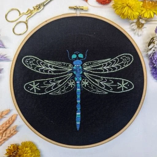 Embroidery hoop with a detailed dragonfly design on black fabric, surrounded by colorful flowers and sewing tools.