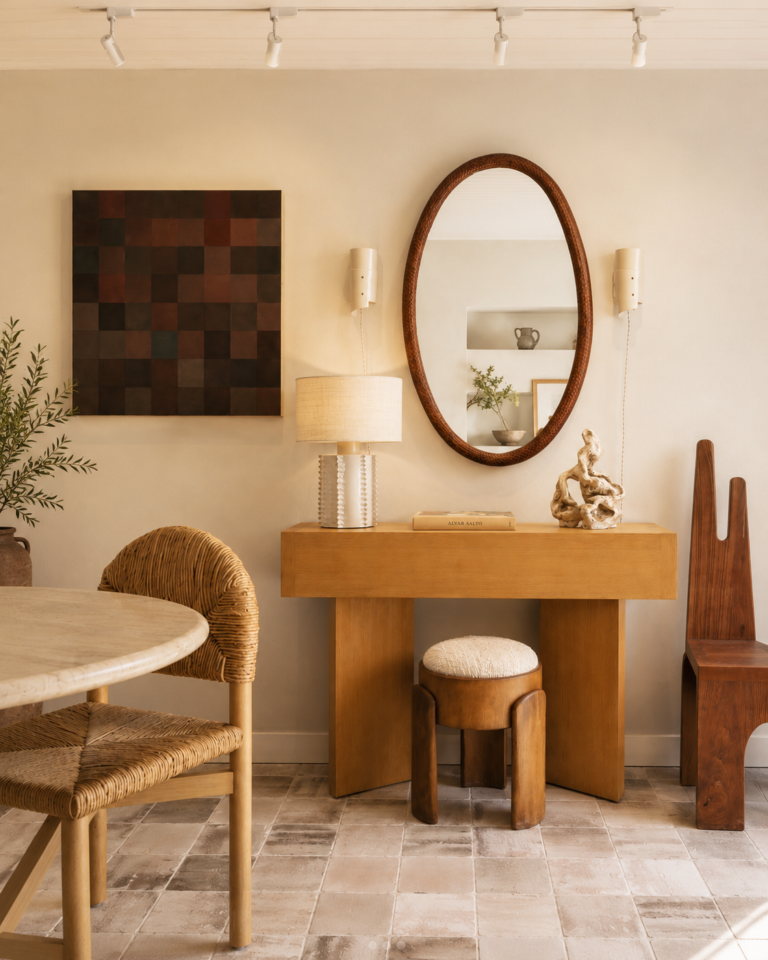 Interior of a room with a wooden console table, a large oval mirror, a table lamp, a sculptural decorative piece, framed pictures, a chair with a woven seat, and a wooden chair, with artwork and plants on the wall.