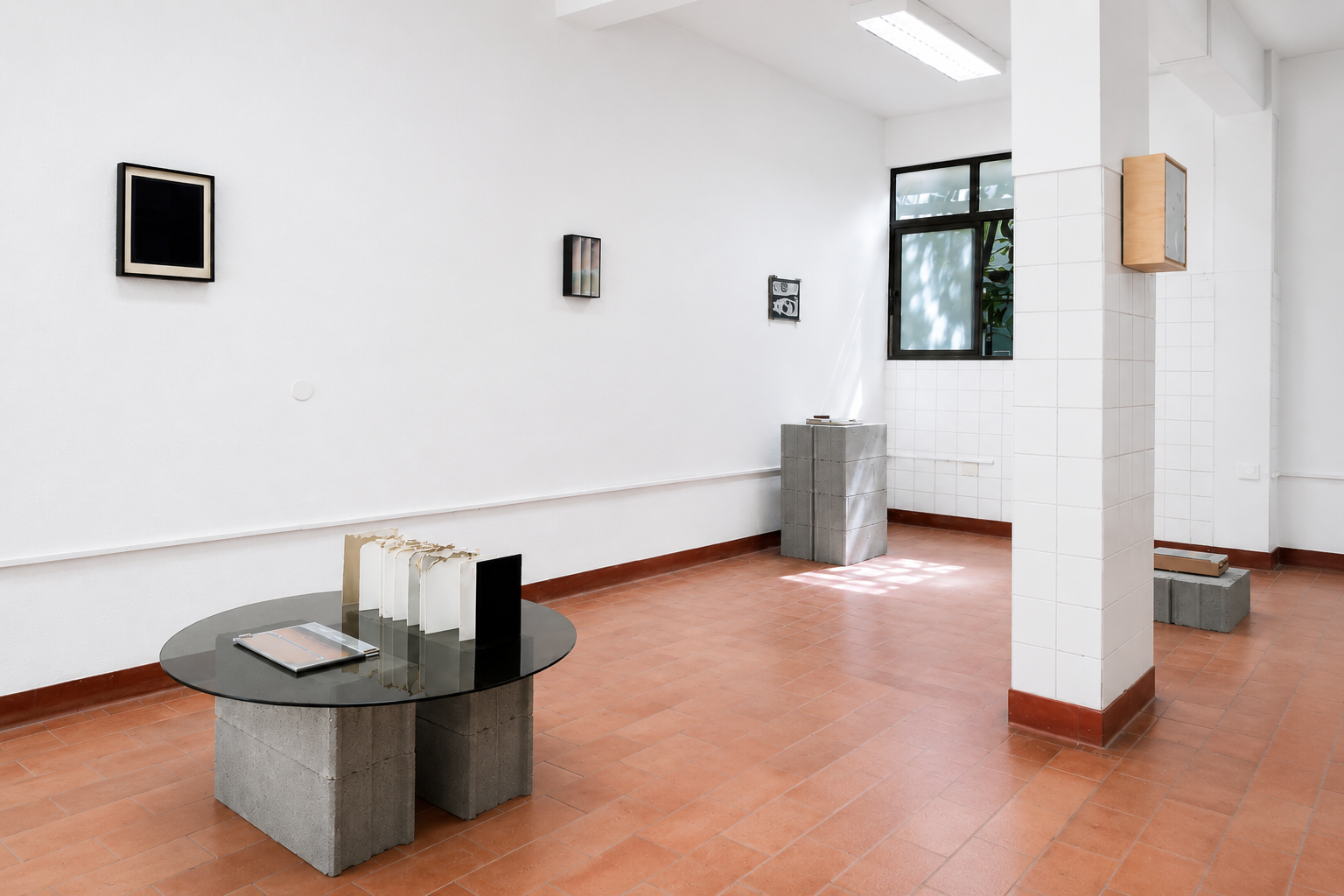 An art gallery with white walls and terracotta tile flooring, featuring small wall-mounted artworks, a round table with books or catalogs, a concrete pedestal, and a window with frosted glass.