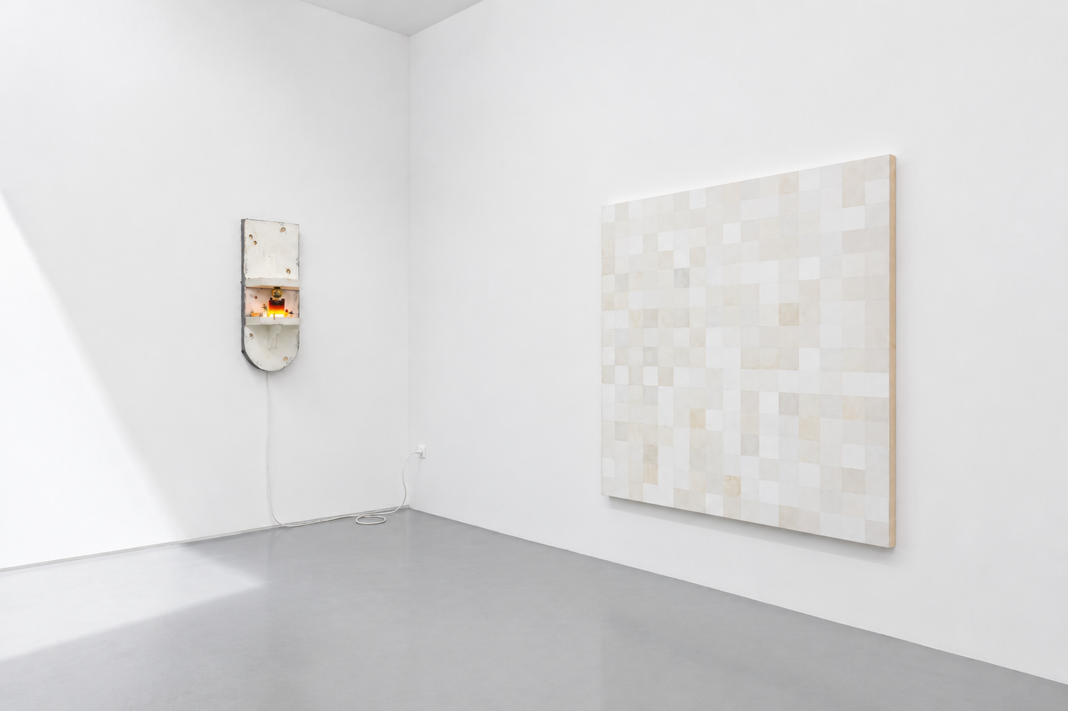 Minimalist art gallery with plain white walls and light gray floor, featuring a small, rustic, white, wall-mounted sculpture on the left and a large square piece of mosaic art with a beige and white pattern on the right.