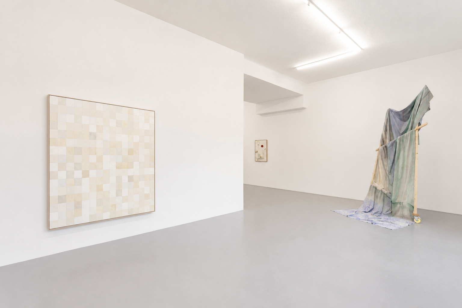 Contemporary art gallery with white walls and grey floor, displaying a pixel art piece on the left, a small mixed media piece on the back wall, and a fabric installation on a wooden frame on the right.