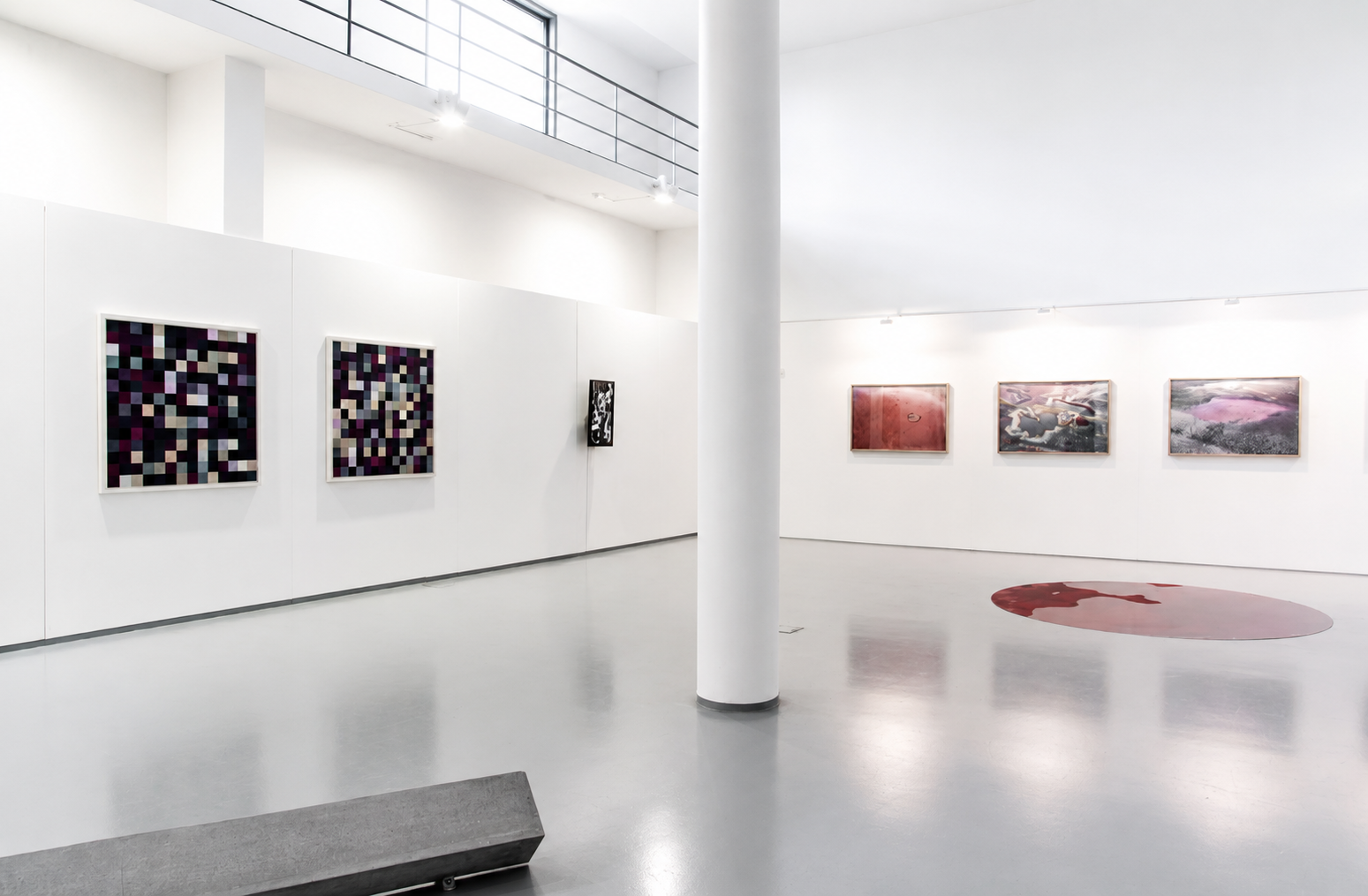 Interior of a modern art gallery with white walls displaying colorful and abstract artworks, including pixelated pieces, black and white designs, and a large floor piece resembling a red and pink abstract shape.