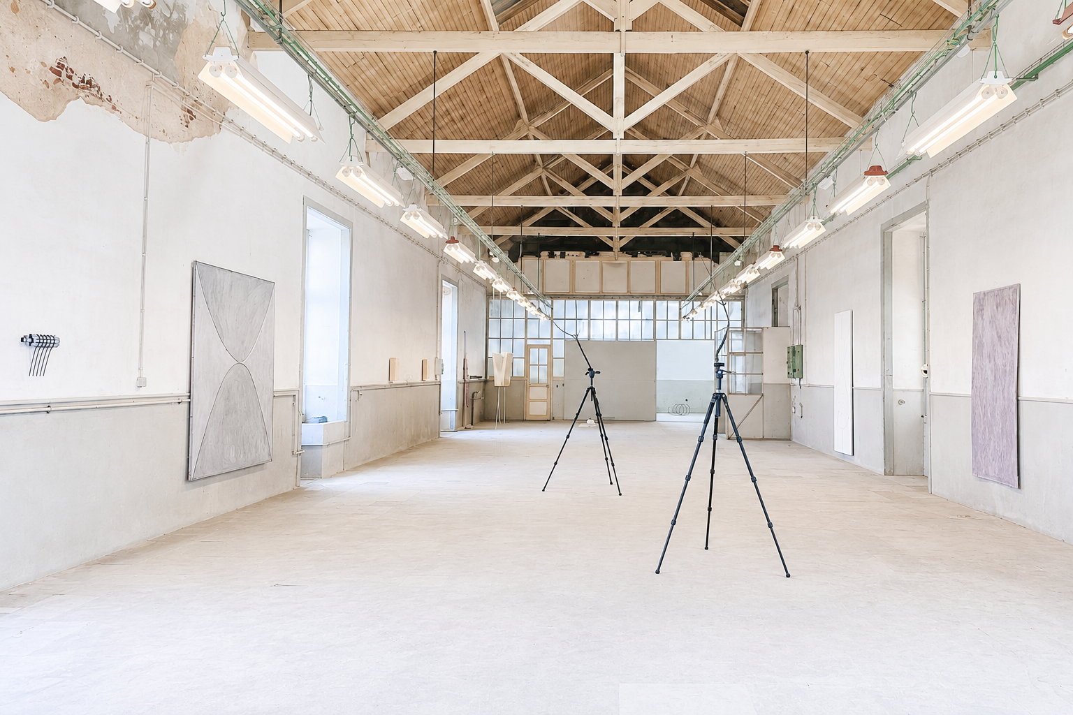 An empty art gallery with white walls, wooden ceiling, and large windows, featuring hanging fluorescent lights and two tripods in the center.