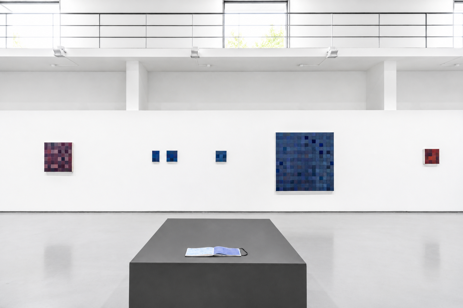 Gallery wall with five pixelated abstract paintings in shades of red, blue, and purple, with an open book on a dark gray pedestal in the foreground.