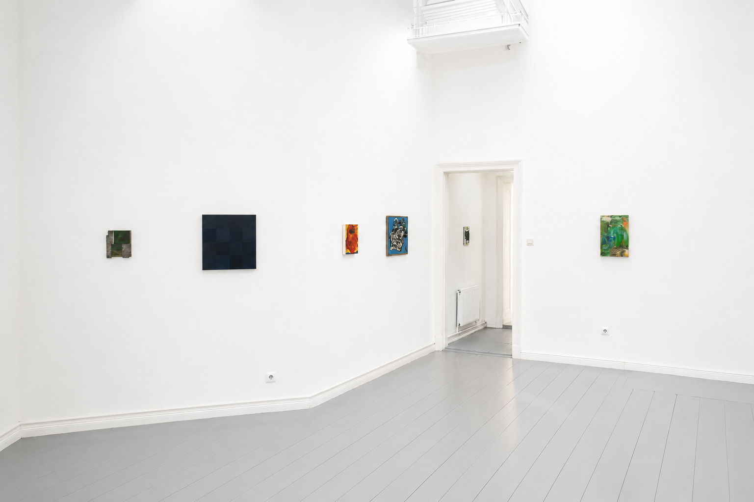 An art gallery with white walls and grey flooring displaying small abstract paintings. A door leads to another room, and there is a white railing on the upper level.