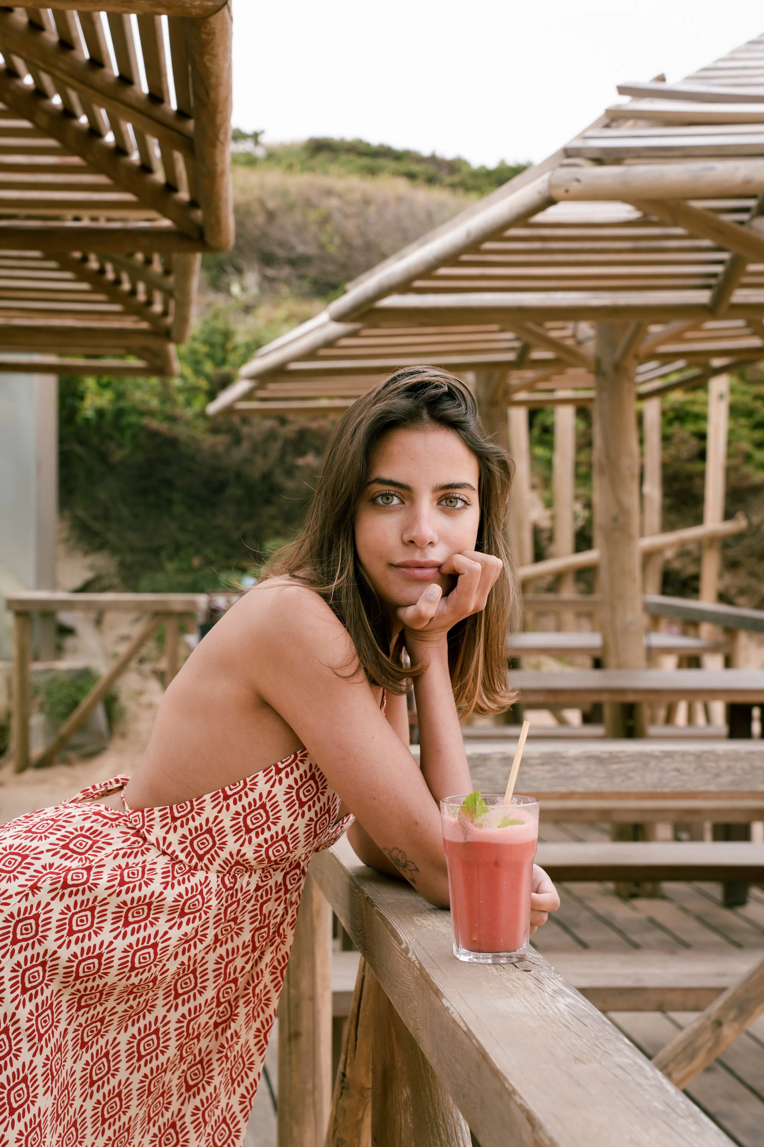 A young woman with long brown hair and blue eyes is sitting at a wooden outdoor bar with a pink cocktail in a tall glass in front of her. She is wearing a red and white patterned skirt and is resting her chin on her hand looking at the camera. In the background, there are wooden structures and lush greenery.