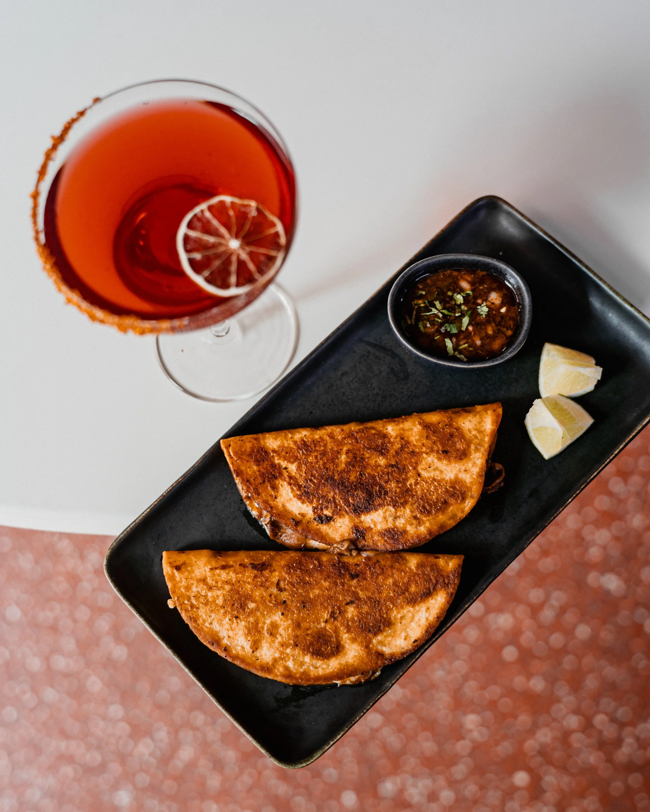 A black rectangular plate with two pieces of toasted quesadilla, slices of lemon, a small bowl of sauce, and a lime wedge. A tall glass of orange-colored beverage with a lemon slice at the top is also visible.