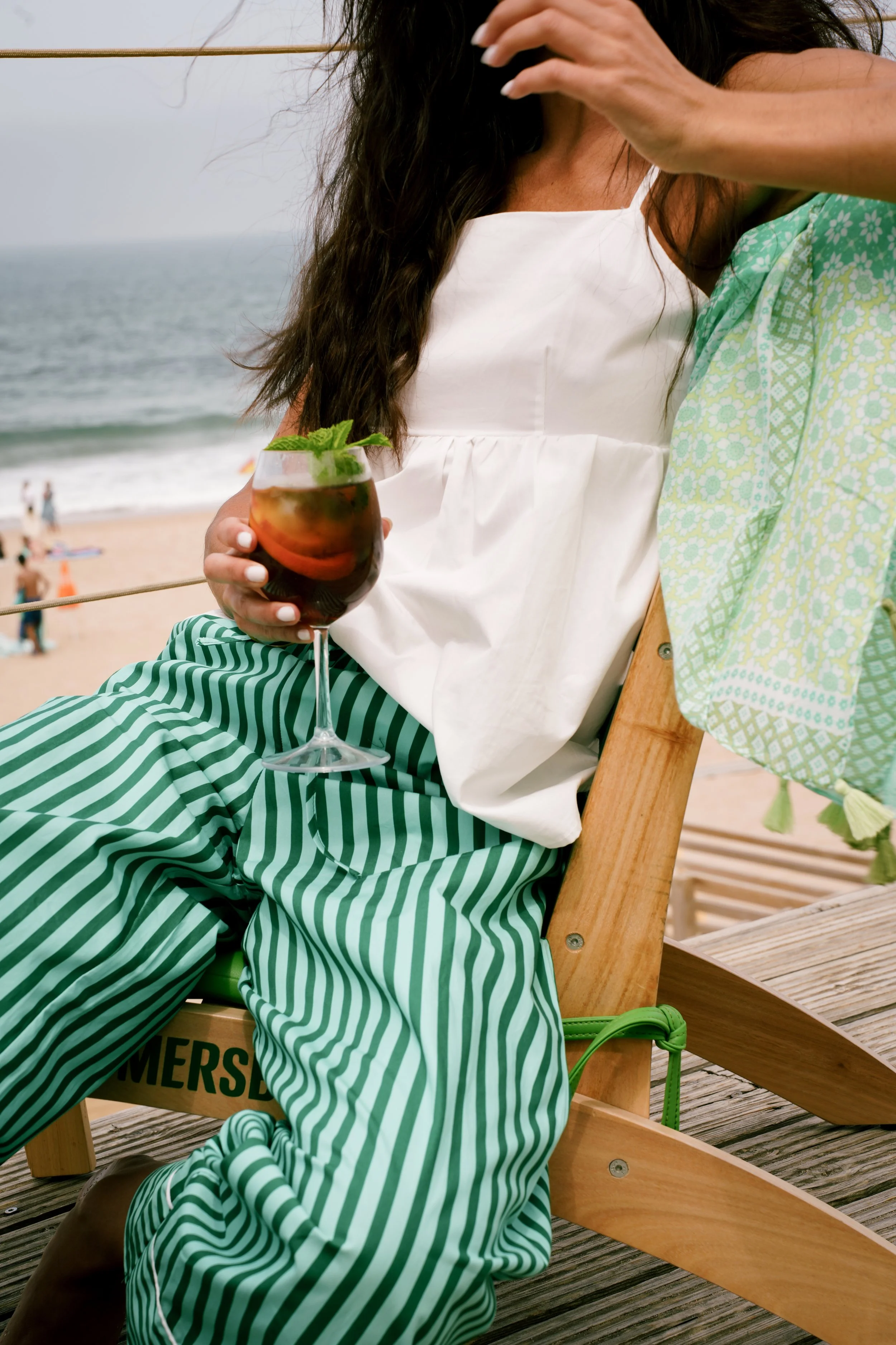 A woman in a white sleeveless top, green and white striped pants, and brown boots, sitting on a wooden chair on a beach, holding a glass of cocktail with mint leaves, with the ocean and beachgoers in the background.