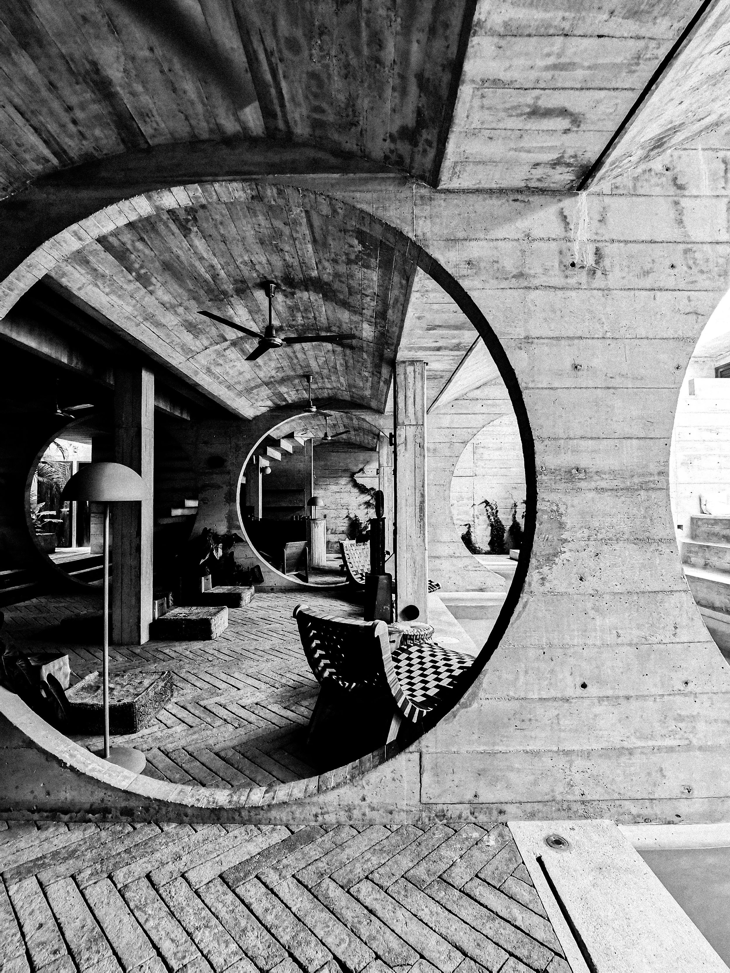 A black and white photograph of an interior space with architectural elements including a large circular mirror, a curved concrete ceiling, wooden flooring, and furniture. The reflection in the mirror shows part of the room with stairs, plants, and hanging lights.