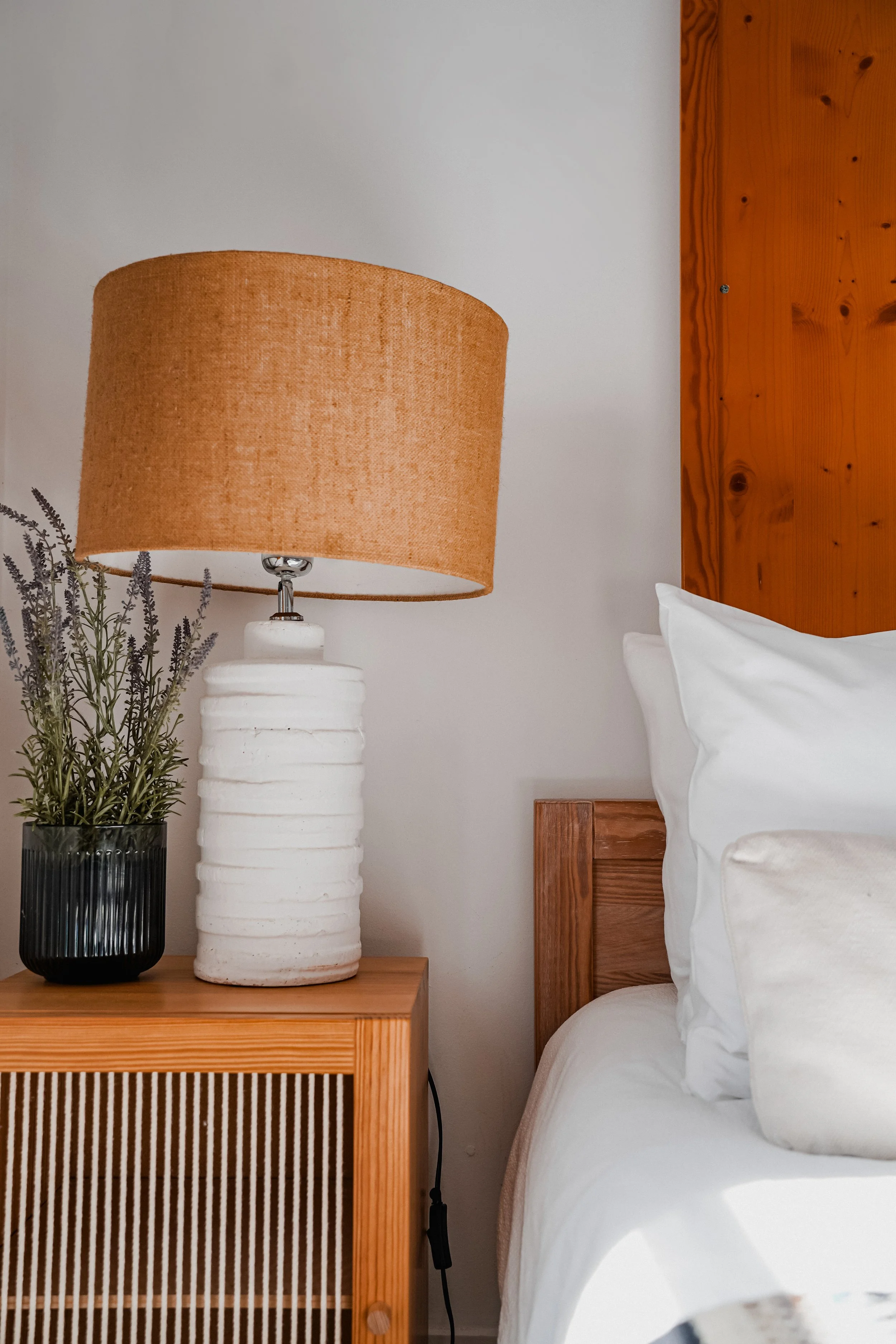 Close-up of a bedside table with a black vase containing lavender and a white ceramic lamp, next to a bed with white bedding and pillows, and a wooden headboard.