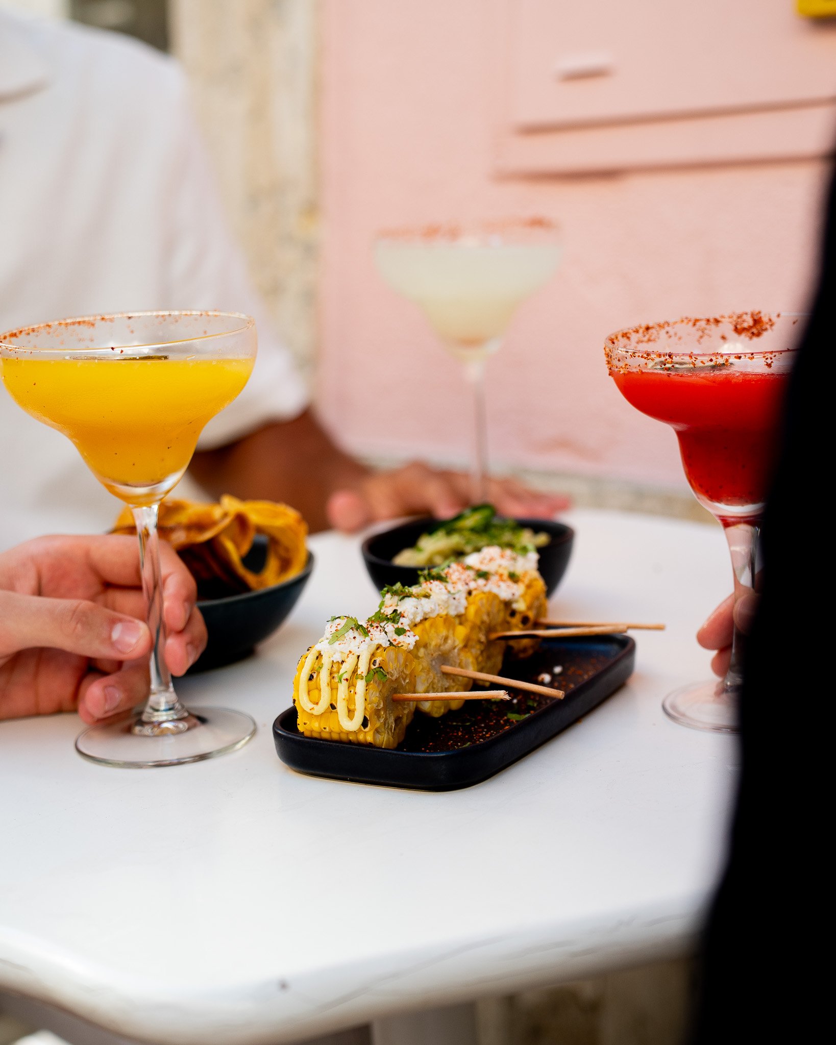 Close-up of a hand holding a yellow cocktail in a glass with a reddish rim, surrounded by three other cocktails of different colors, along with sushi rolls and bowls of food on a white table.