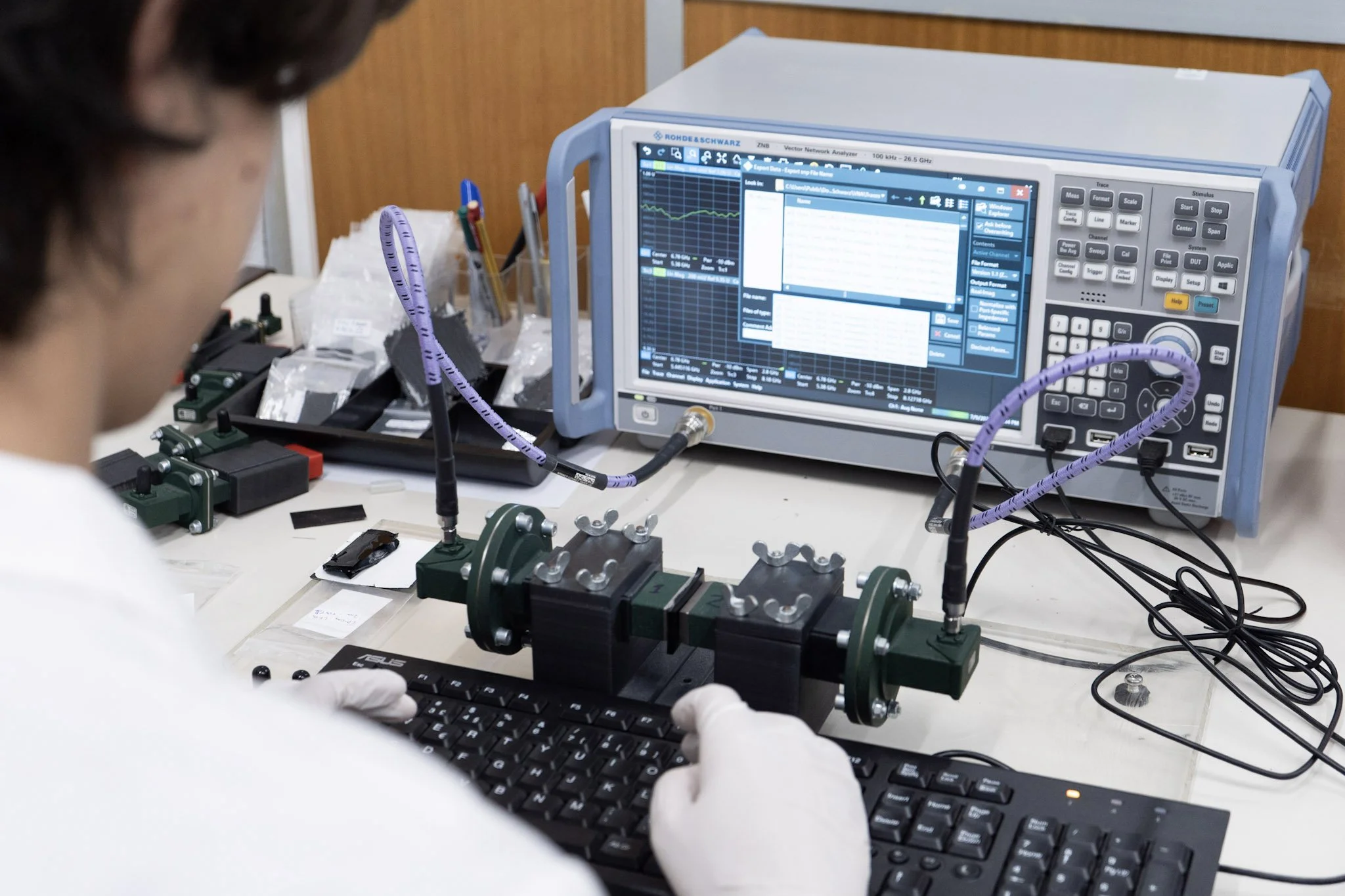 A scientist working at a lab bench with electronic testing equipment, including a Rohde & Schwarz network analyzer, connected to various circuit components and a computer keyboard.