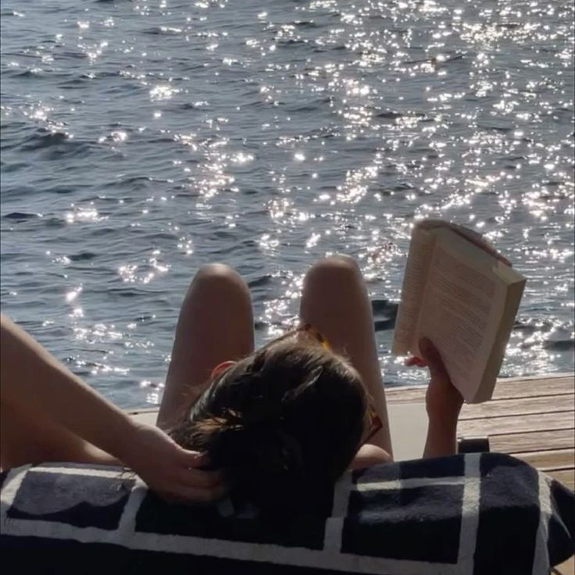 Person relaxing on a lounge chair at the beach, holding a book, with the ocean view in the background and sunlight reflecting on the water.